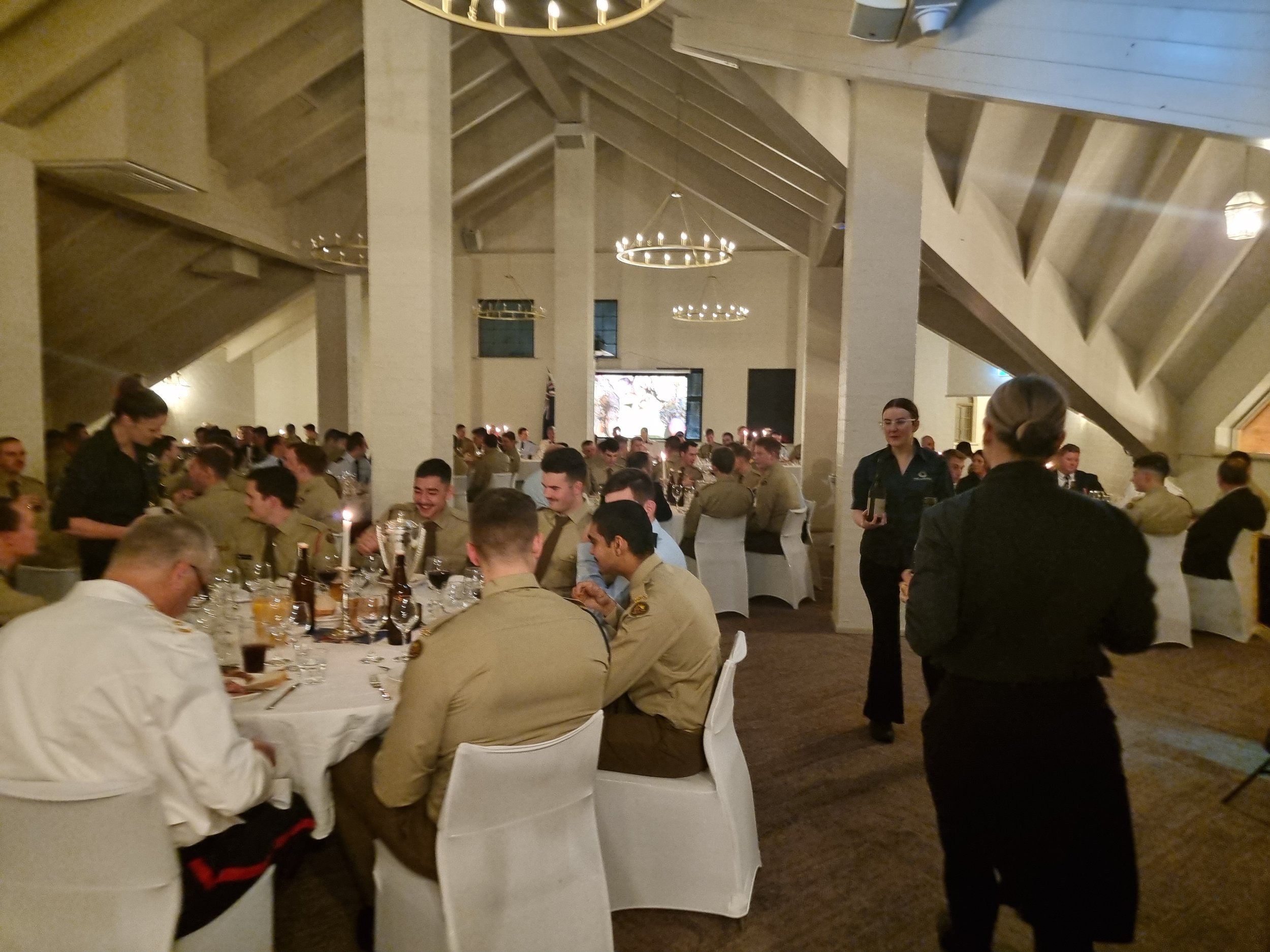 A formal dinner event with military personnel seated at round tables in a large decorated hall, with waitstaff serving drinks.