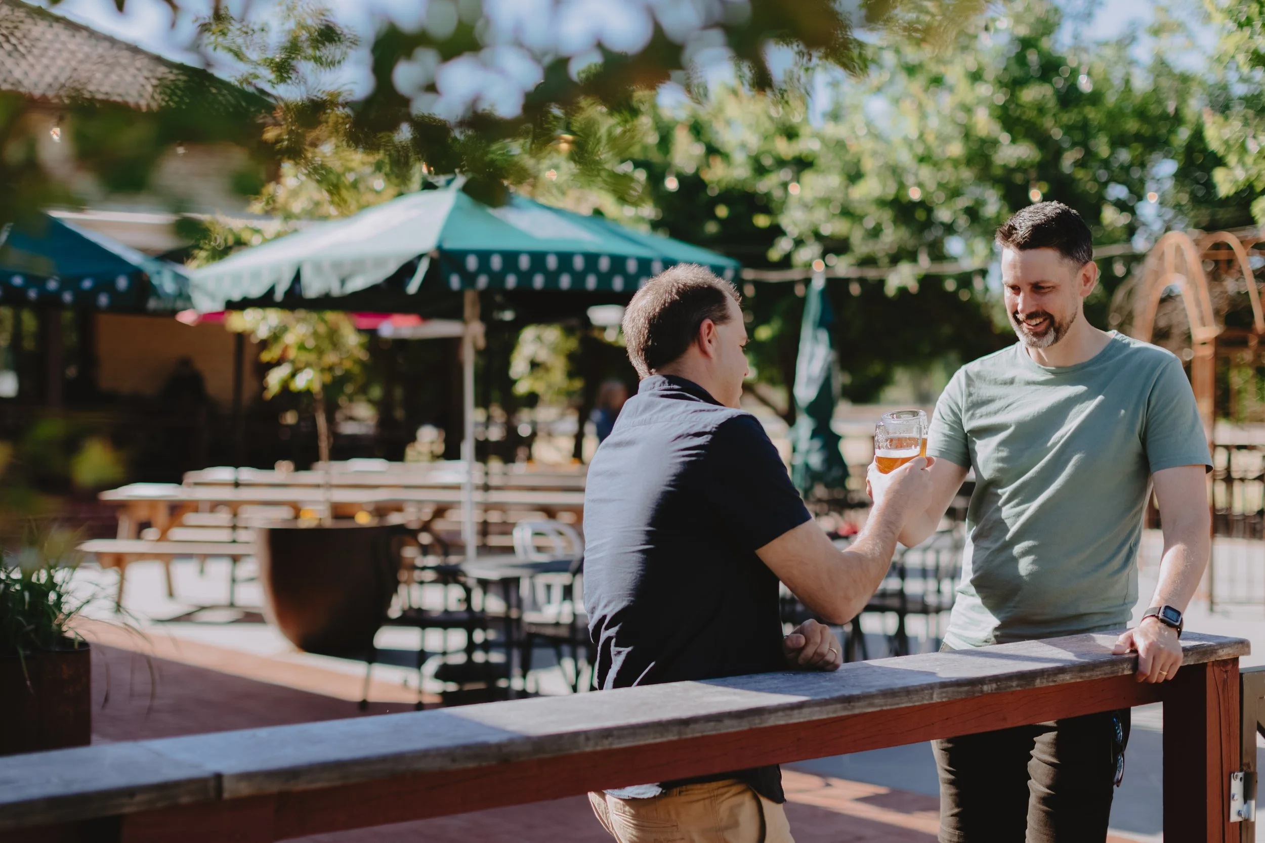 Two men engaging in a toast at an outdoor patio, with chairs, tables, and umbrellas in the background, surrounded by green trees.