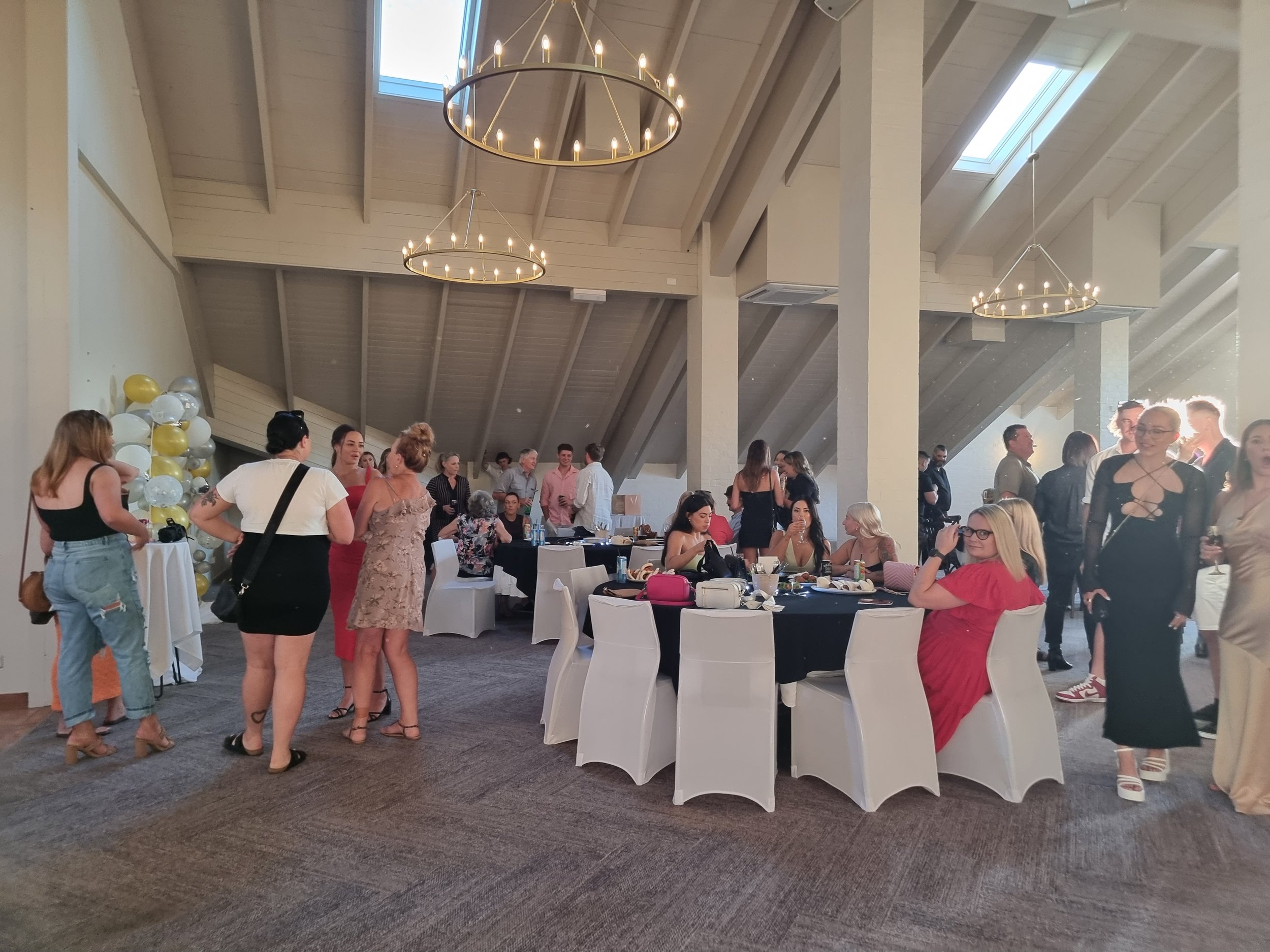 People socializing at a party, sitting and standing around tables, with balloons on the left, and chandeliers hanging from a high ceiling with skylights.