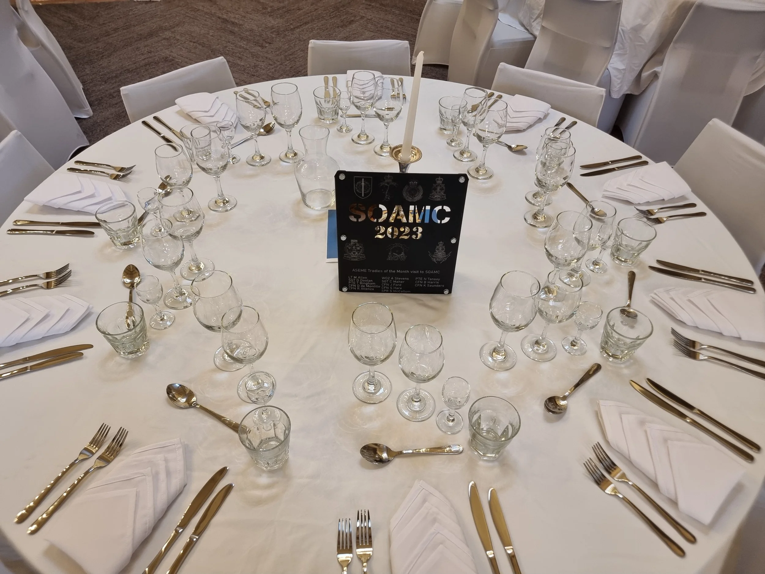 Round banquet table set with white tablecloth, white napkins, gold utensils, and various glasses, with a black SOAMIC 2023 event sign in the center.