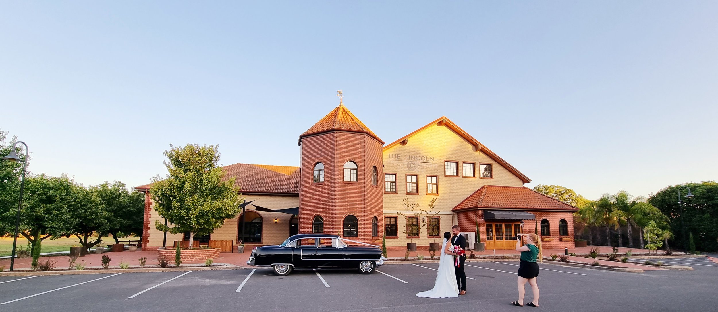A wedding couple standing in front of a vintage black car outside a brick and beige building with a tower, trees, and a parking lot.