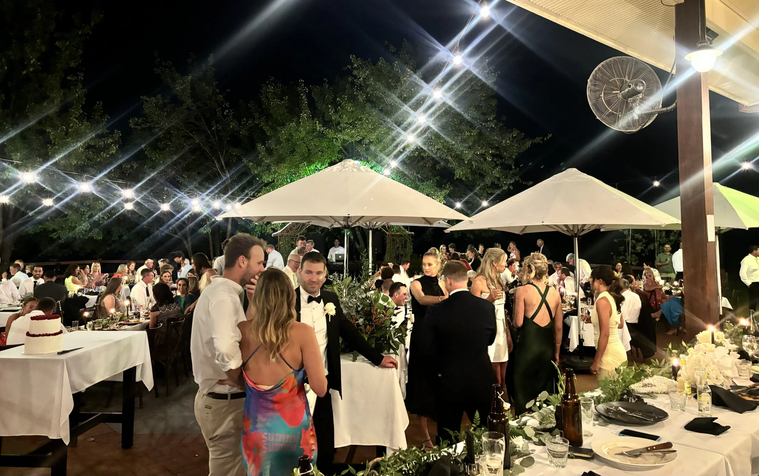 Nighttime outdoor wedding reception with guests socializing, elegantly dressed, under string lights, with tables decorated with floral arrangements and food.