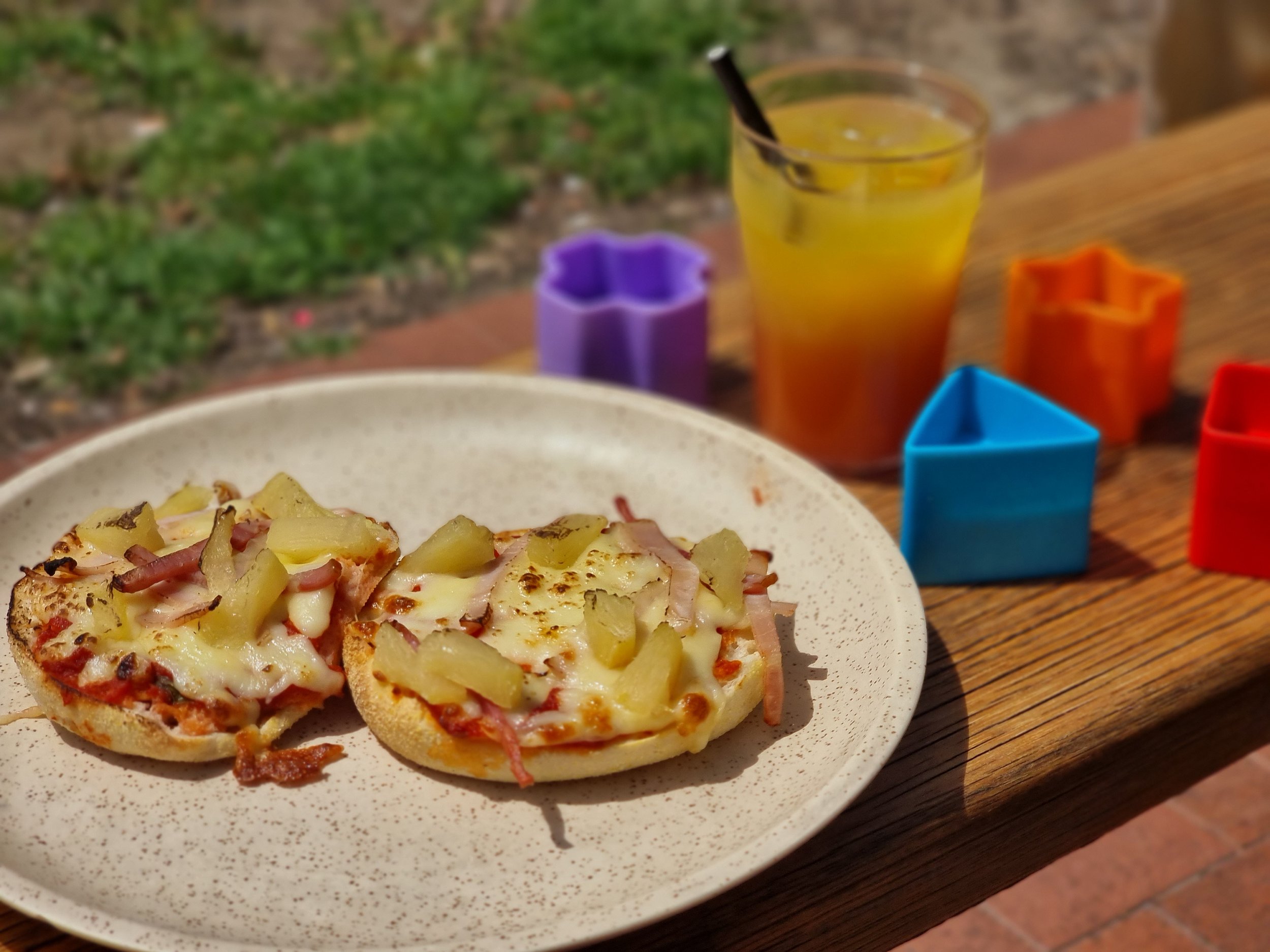 Two pineapple and ham personal pizzas on a beige plate, a glass of yellow-orange drink with a straw, and colorful cookie cutters on a wooden table outdoors.
