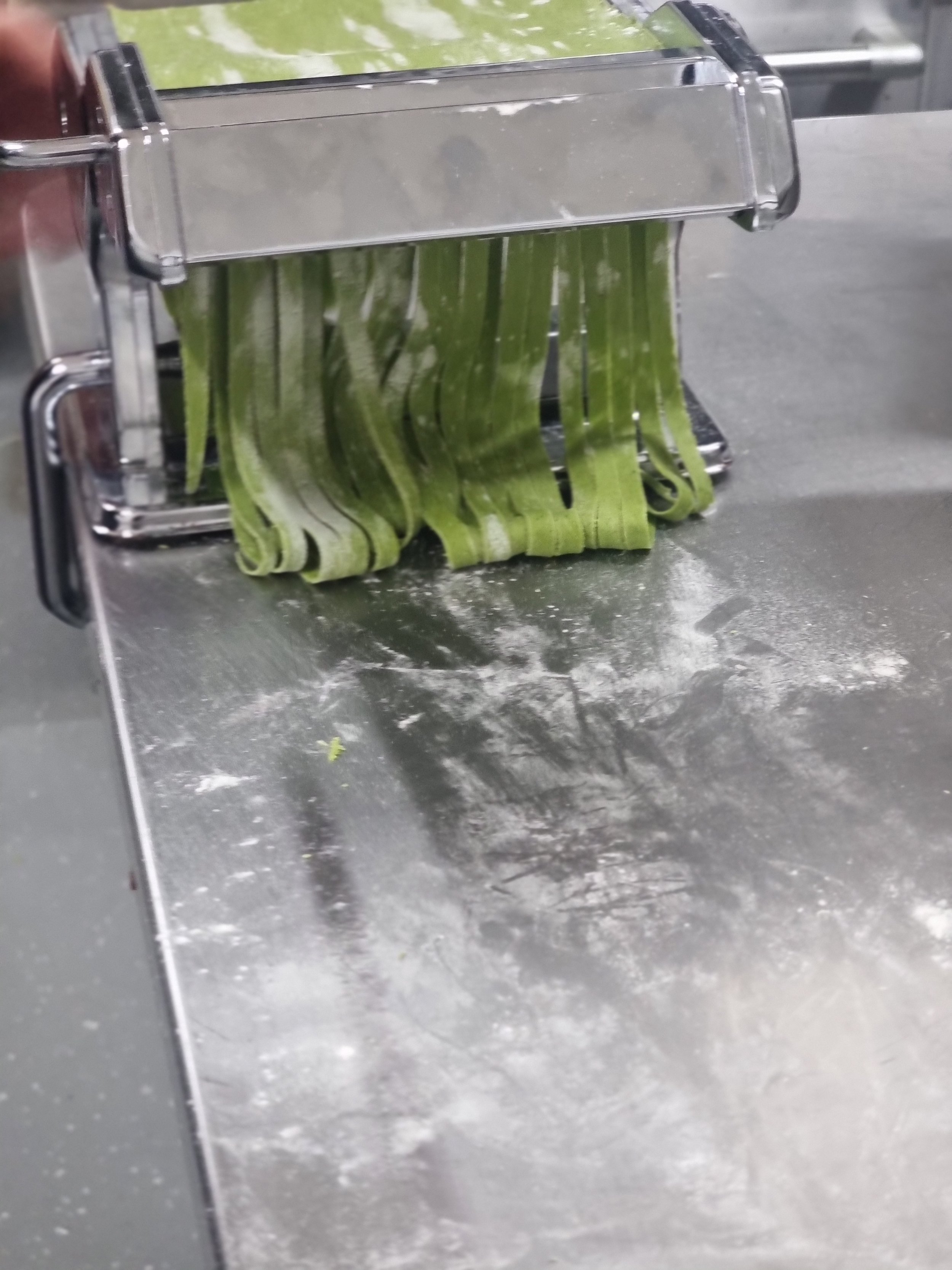 Fresh green spinach pasta being made with a pasta machine on a floured metal surface.
