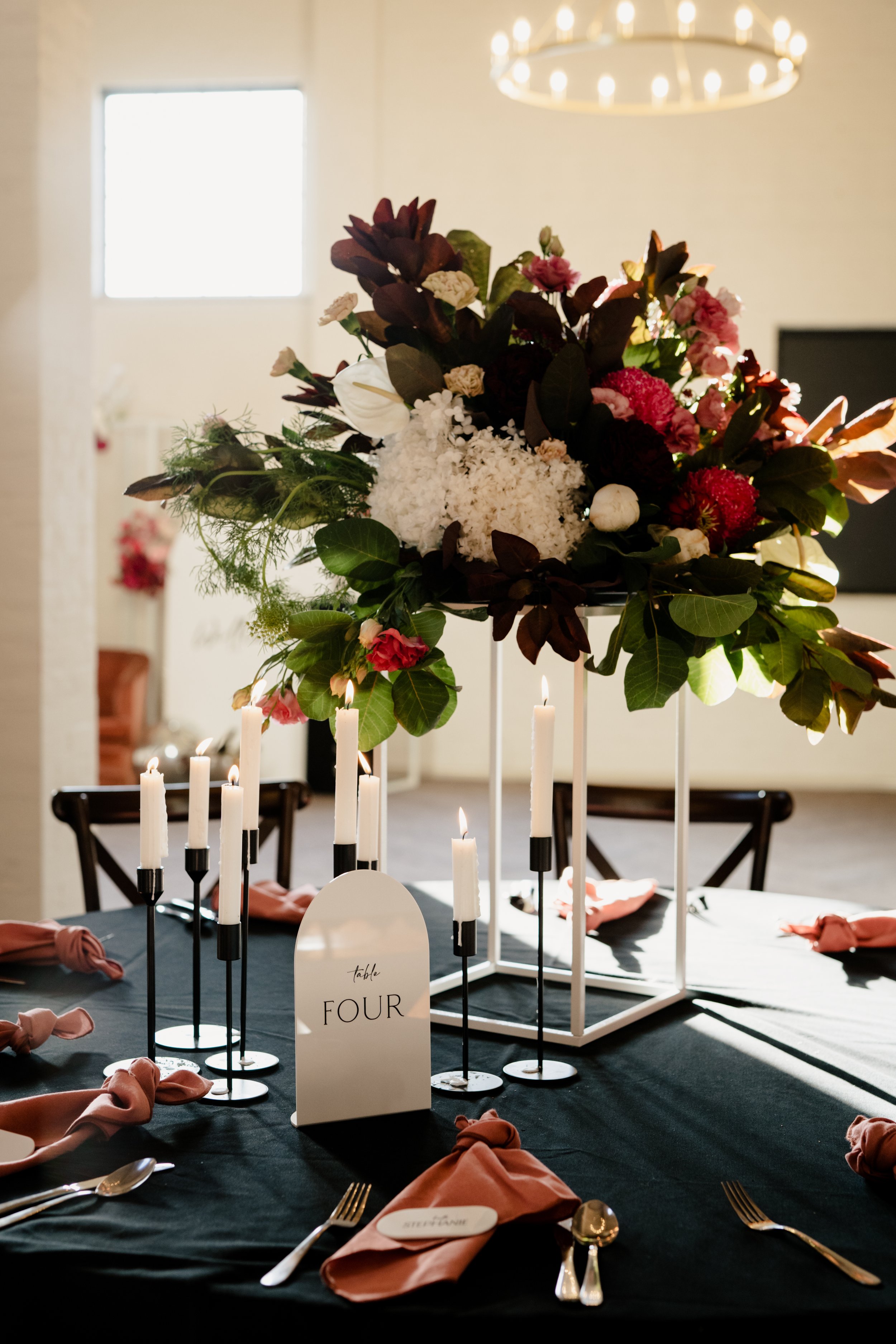 Elegant dining table with black tablecloth, pink napkins, and gold cutlery, decorated with tall white candles and a large floral centerpiece of white, pink, and dark foliage, with a table number card labeled 'FOUR' at a formal event.