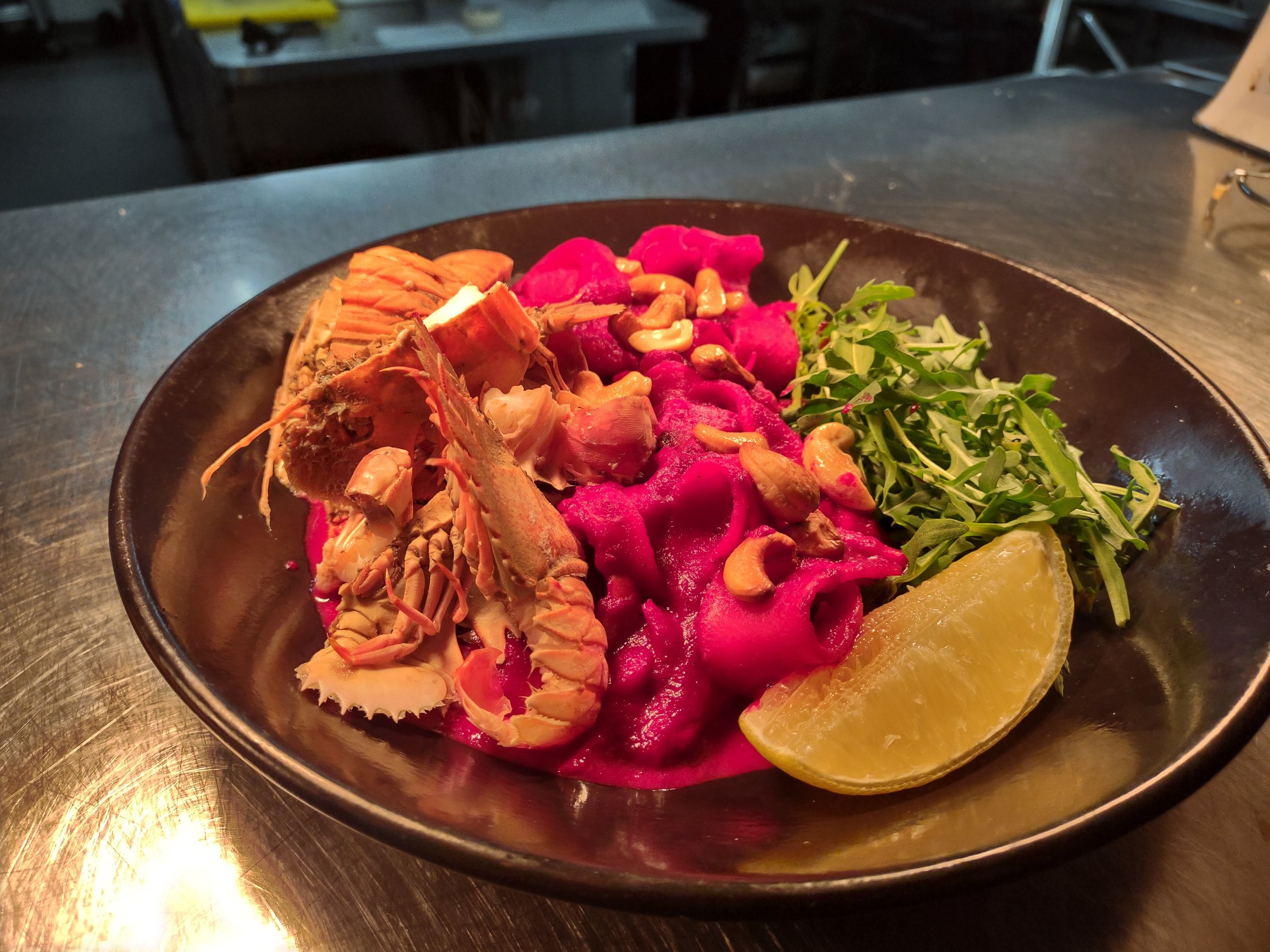 A black bowl containing vibrant pink pasta topped with lobster, garnished with fresh greens, chopped nuts, and a lemon wedge, on a dark metal table.