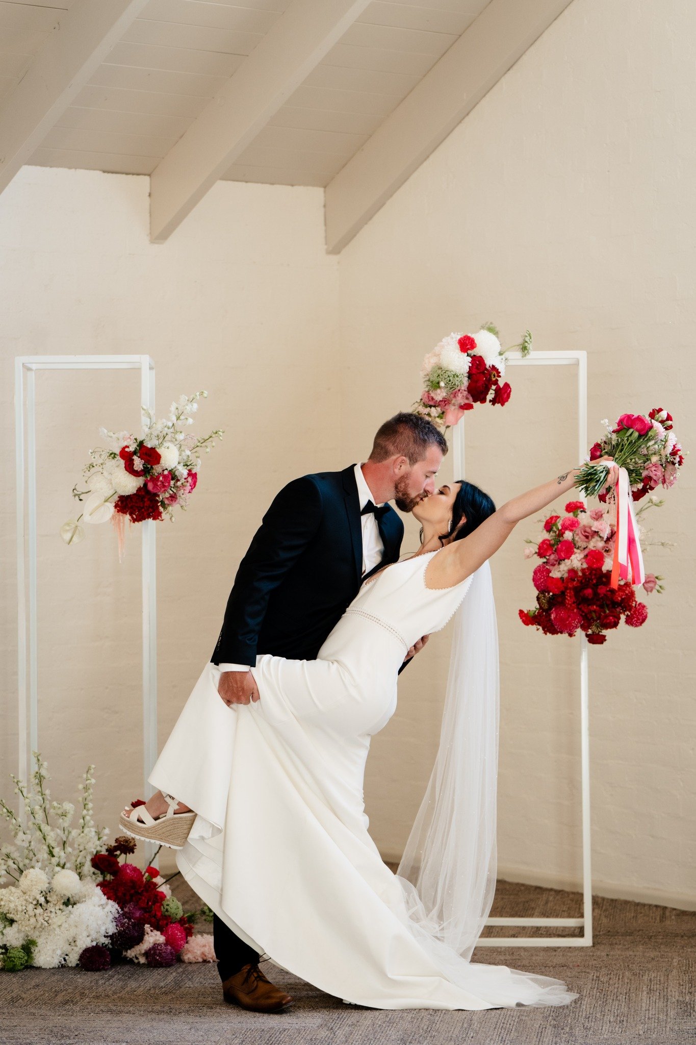 A newlywed couple sharing a kiss during their wedding ceremony. The groom, in a black tuxedo, is dipping the bride, in a white dress, who has her arms raised holding two bouquets of pink and red flowers. There are floral arrangements on white geometr