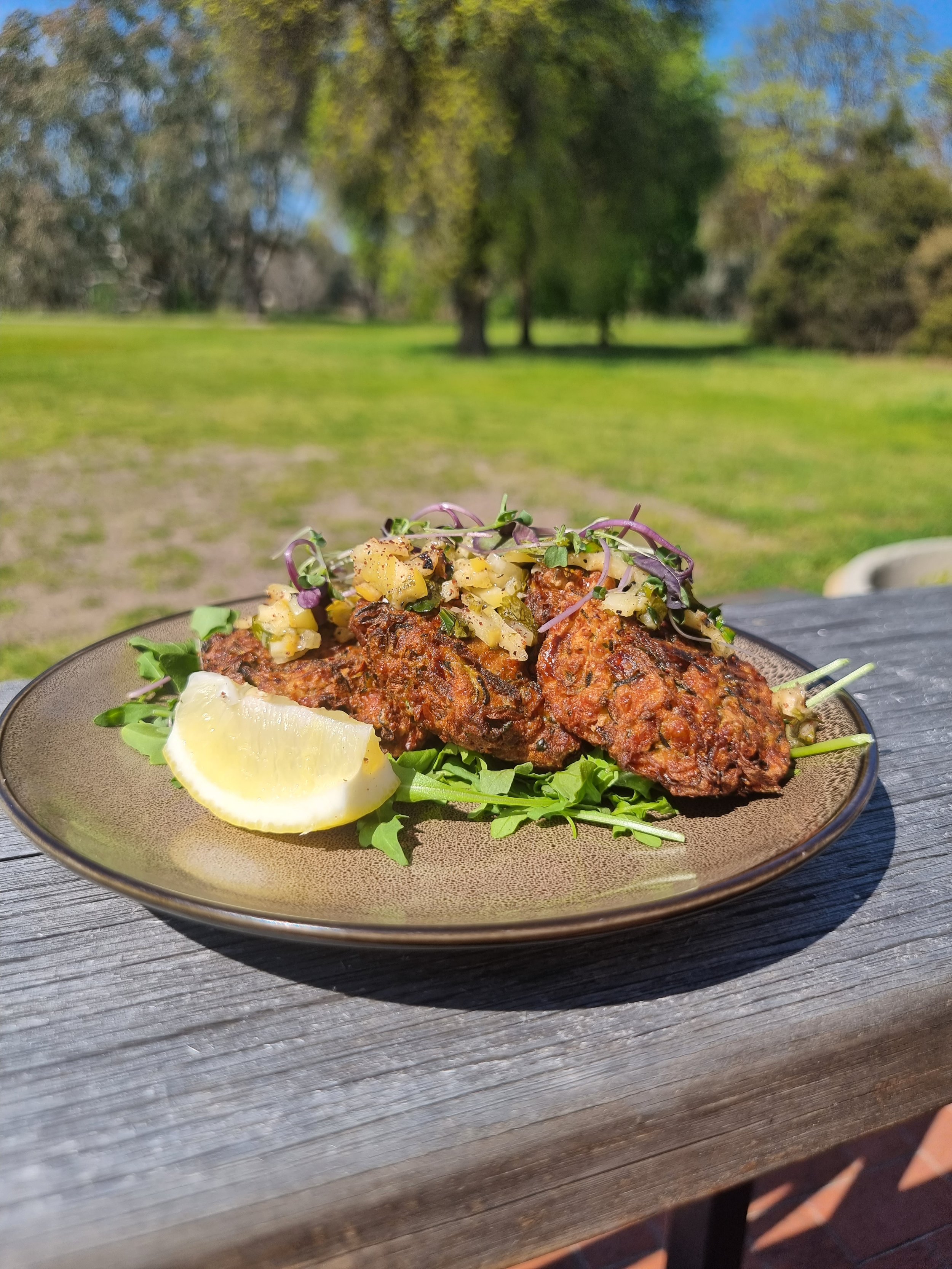 Plate of three skewered grilled meat pieces with a lemon wedge and a topping of diced vegetables, set on a bed of greens outdoors on a wooden table with a grassy park and trees in the background.