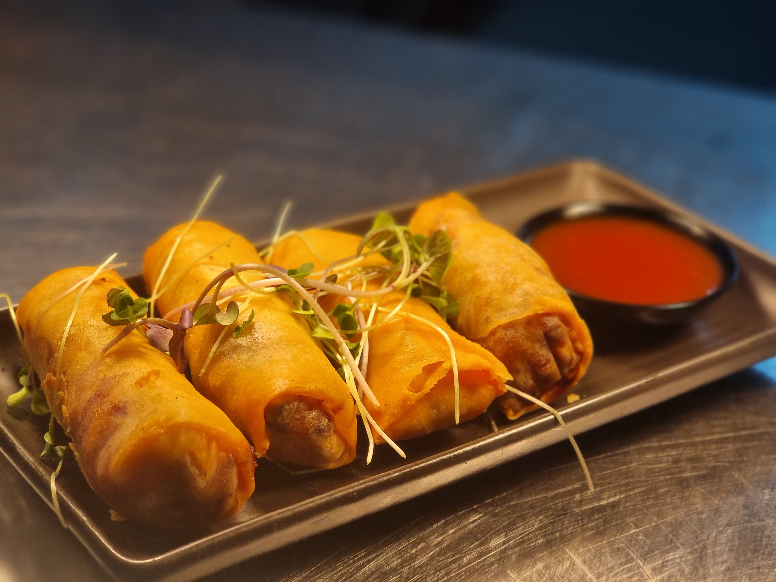 Four spring rolls garnished with sprouts on a metal tray with a side of red dipping sauce.