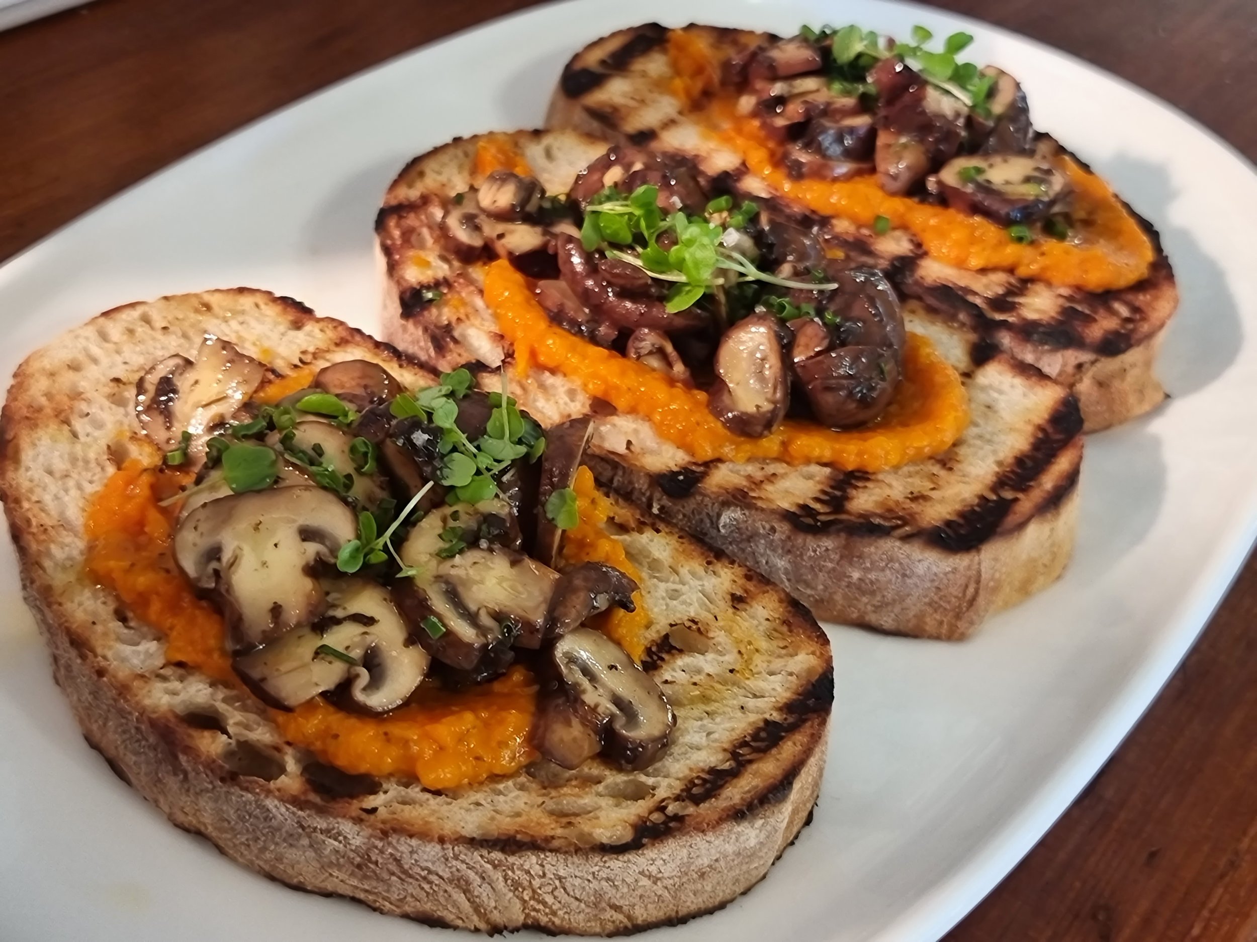 Grilled bread topped with mushroom, roasted red pepper, and microgreens on a white plate.