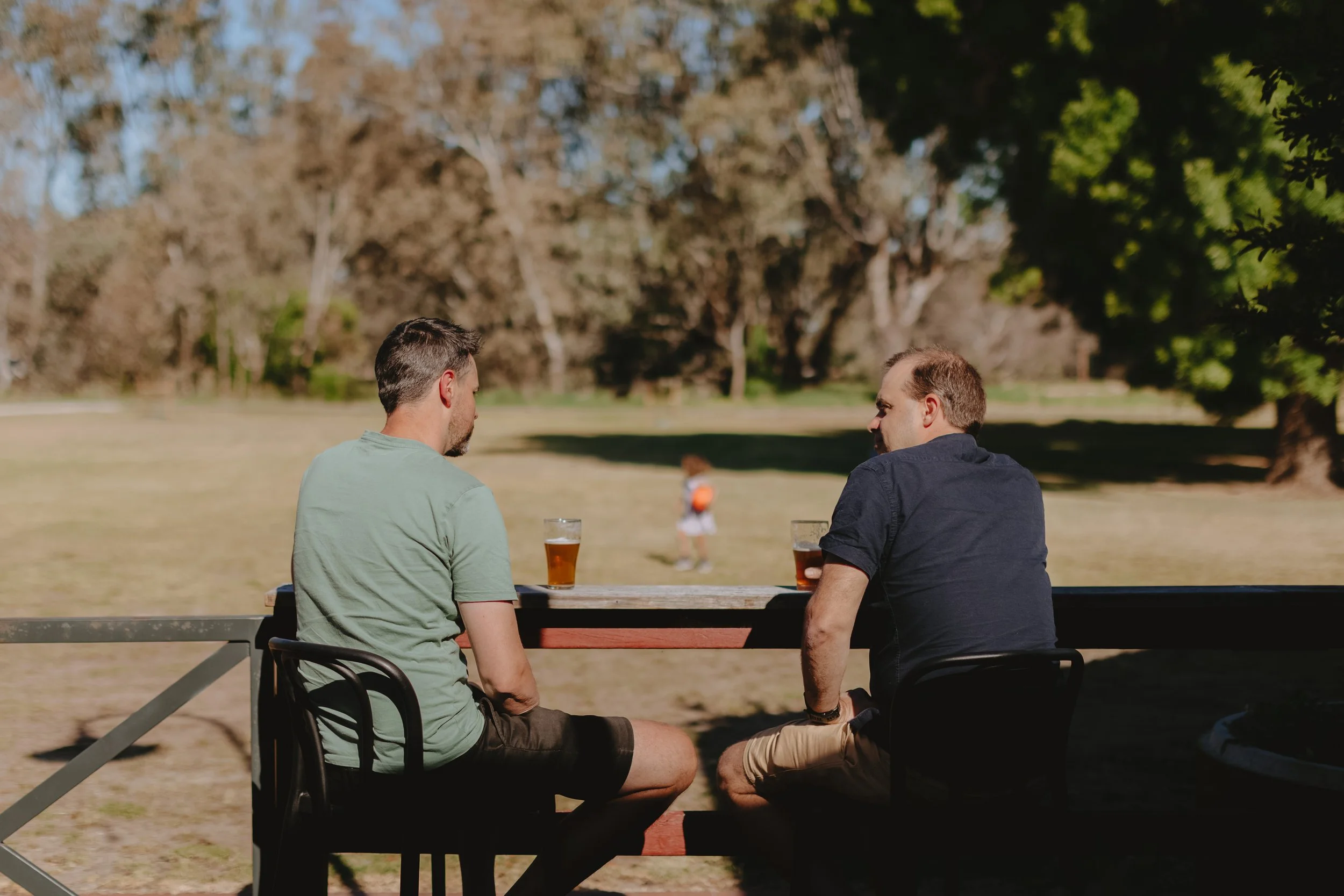 Two men sit at an outdoor table with drinks, facing a grassy park with trees, while a woman walking in the background.