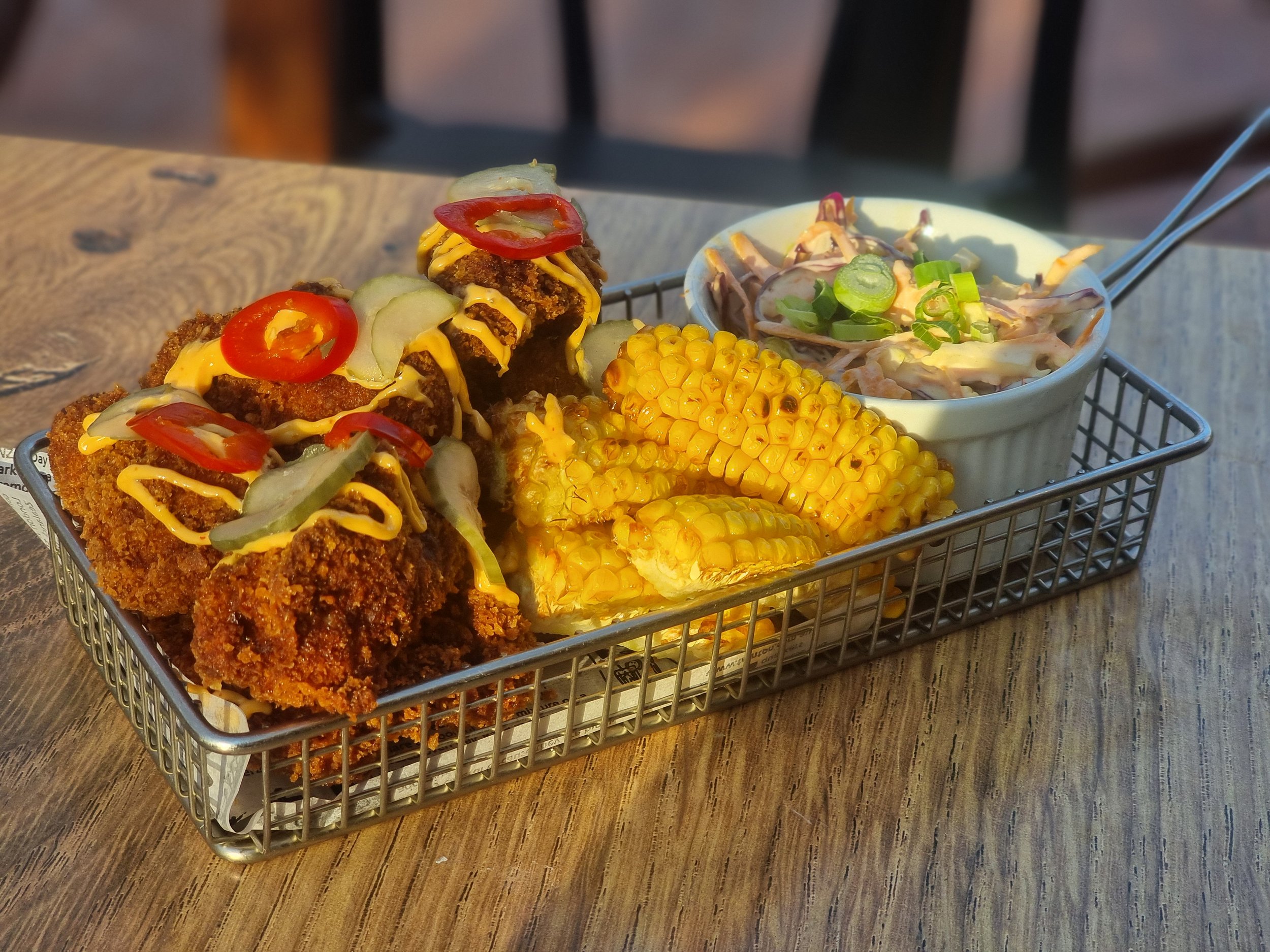 Fried chicken tenders with mustard, pickles, and hot peppers, grilled corn on the cob, and coleslaw in a small bowl on a metal tray.