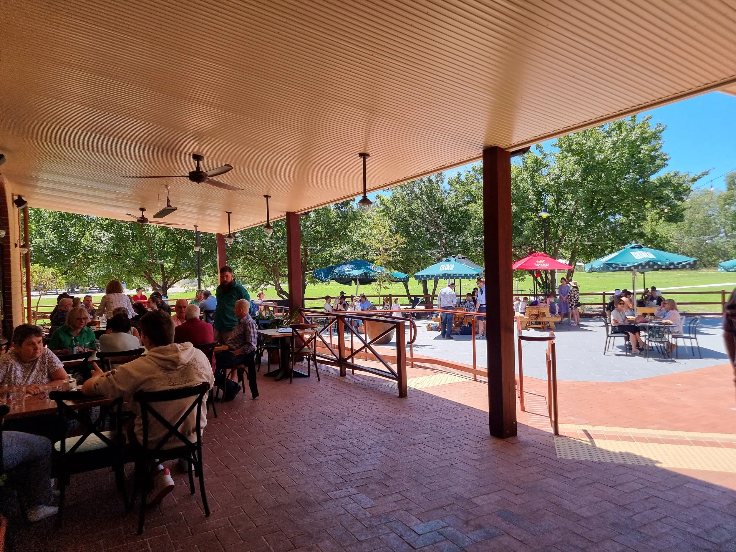 People dining inside a shaded patio area and outside at tables under umbrellas at a park.