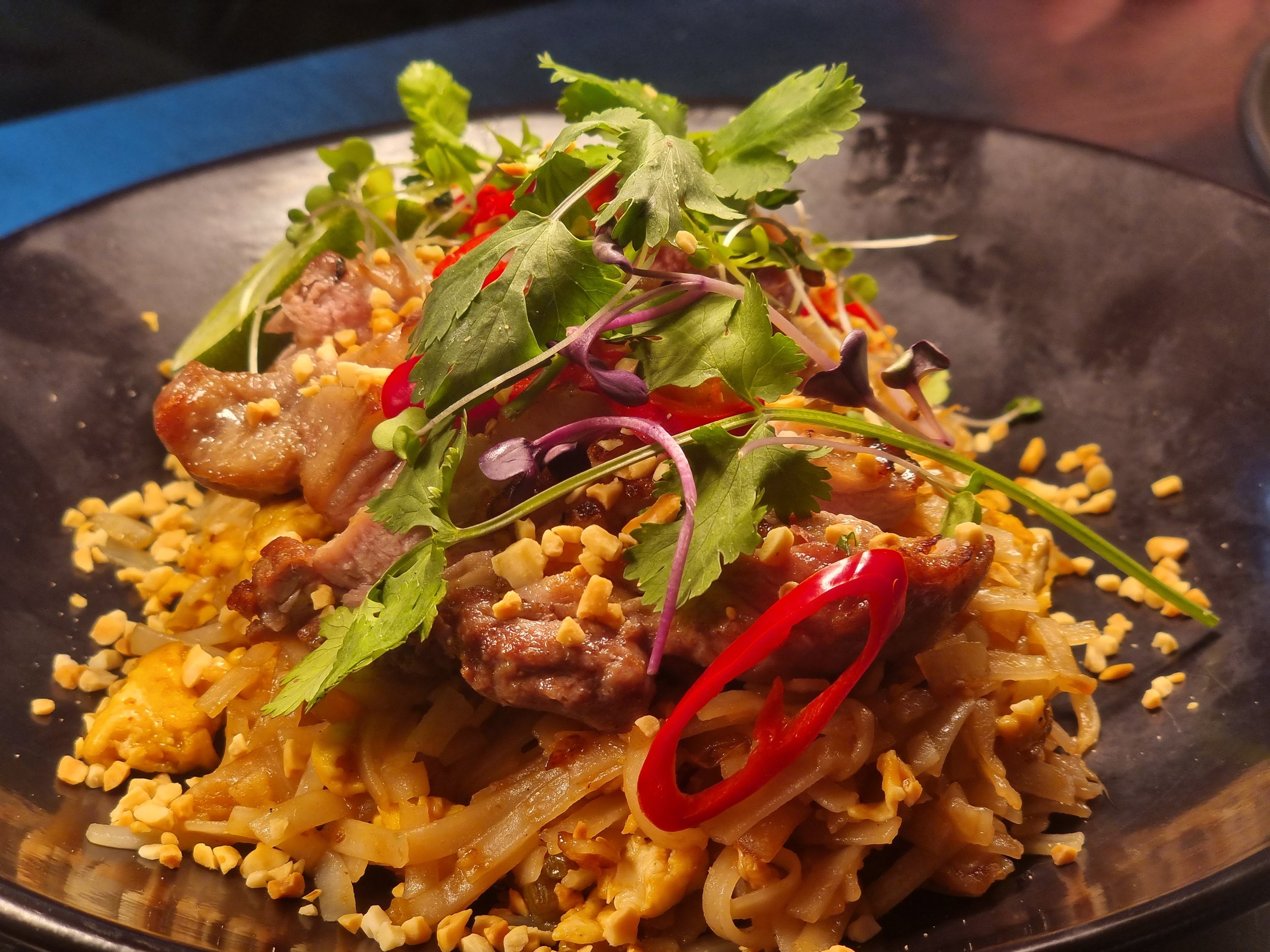 A dish of noodles topped with cooked meat, chopped peanuts, fresh herbs, and sliced red chili peppers.