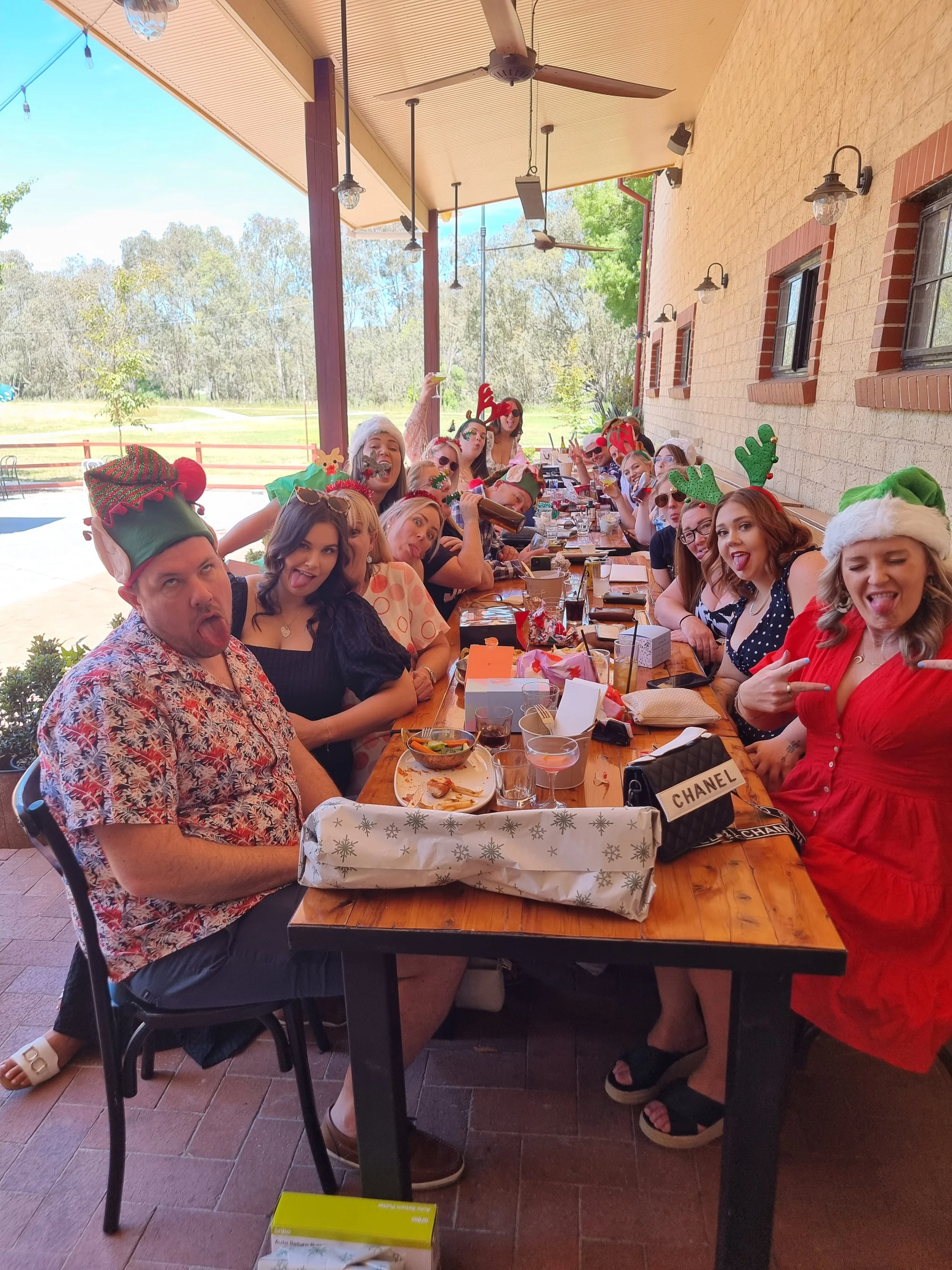 Group of people at a Christmas party wearing holiday hats and accessories, sitting at a long table outdoors on a covered patio, with some making playful faces.