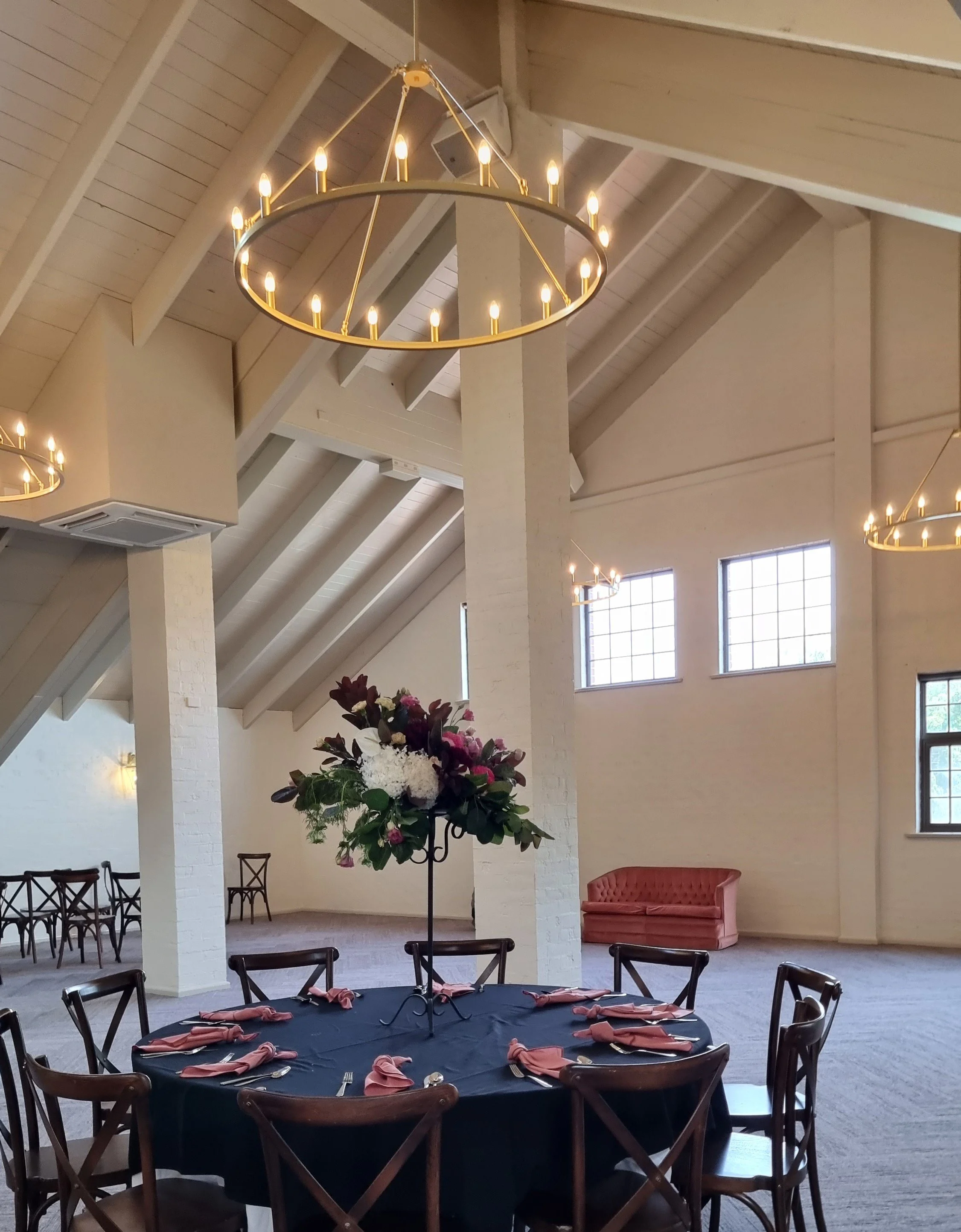 Elegant event space with high vaulted ceilings, chandeliers, a round table with a floral centerpiece, navy tablecloth, pink napkins, and chairs, with ample natural light from multiple windows.