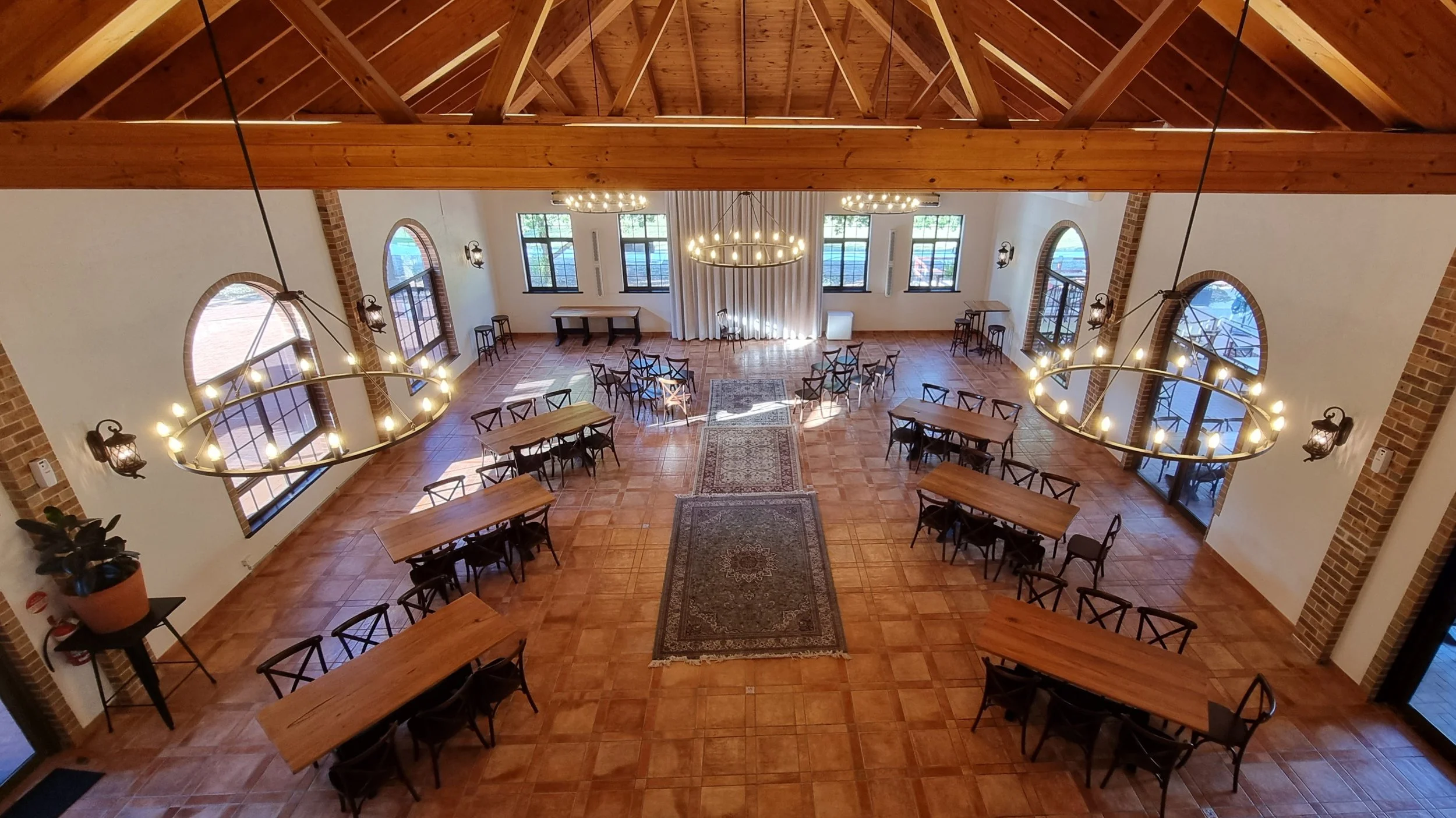 A spacious event hall with wooden ceiling beams, large arched windows, and a tiled floor. The room is decorated with round chandeliers and wall sconces, with tables and chairs arranged around the space. A patterned rug runs down the center, leading t