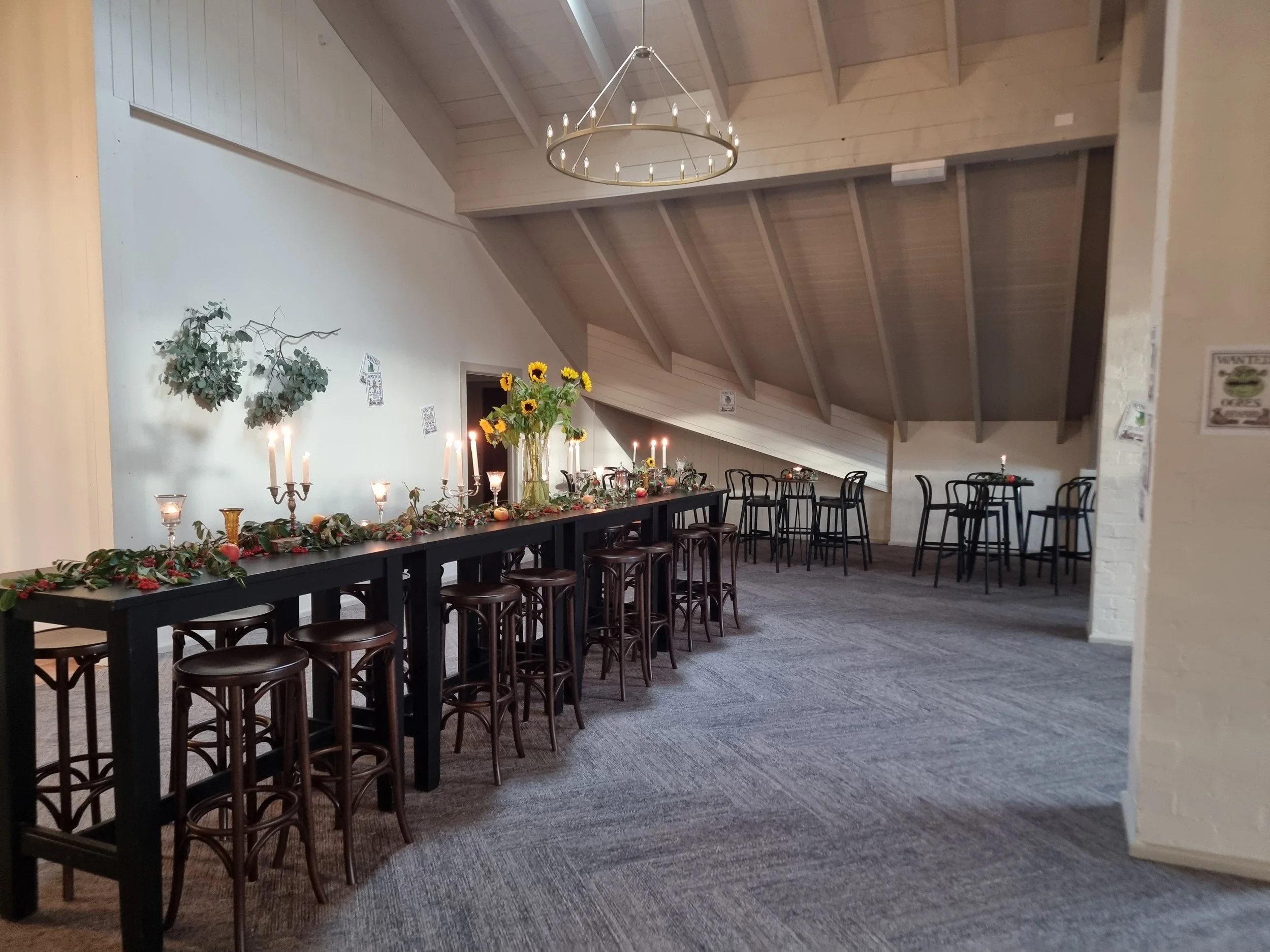 Decorated indoor event space with a long table adorned with candles, greenery, and sunflowers, and several smaller tables with chairs in a room with sloped ceiling and light-colored walls.