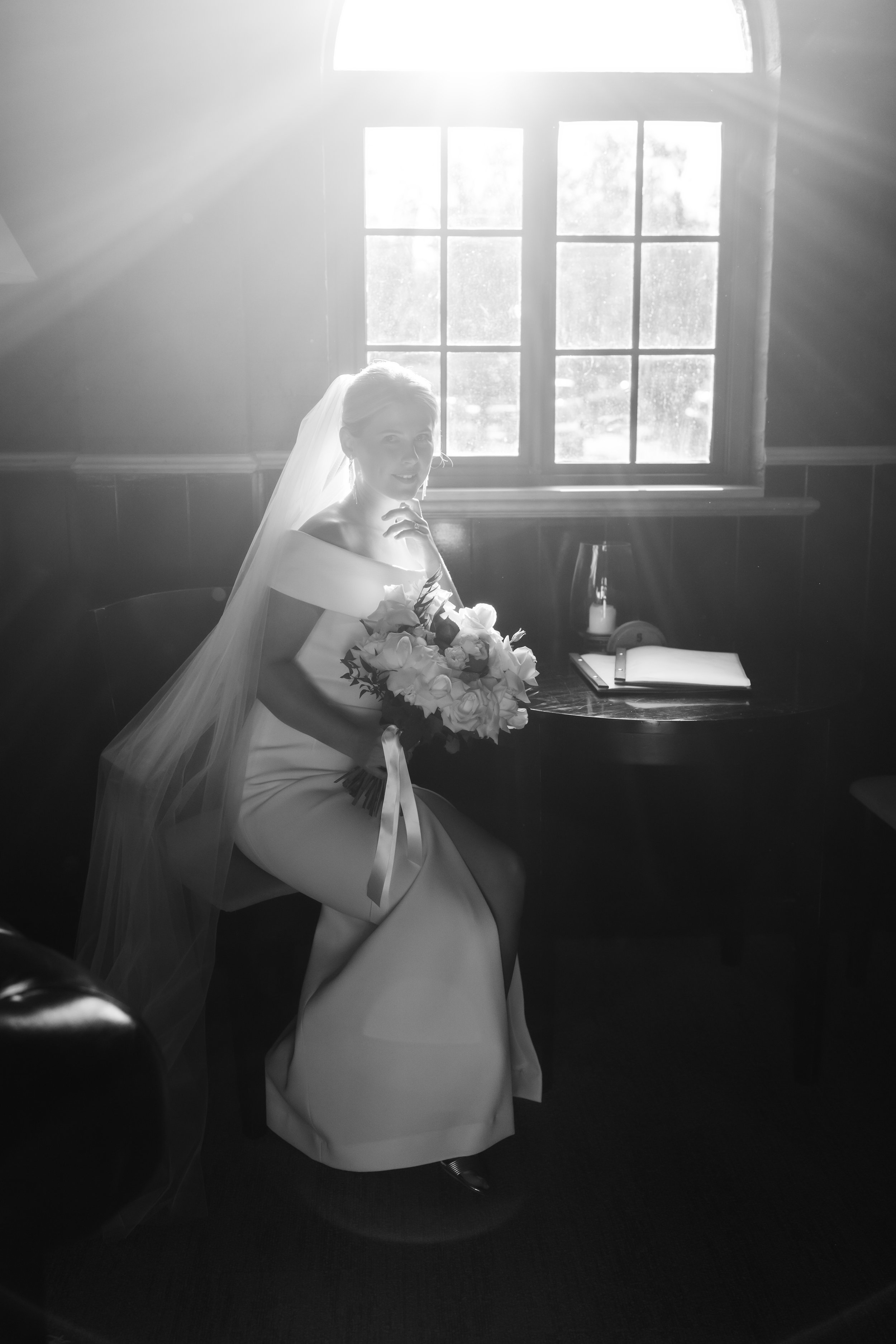 A bride sitting on a chair in front of a window, holding a bouquet of flowers, with sunlight streaming in through the window.