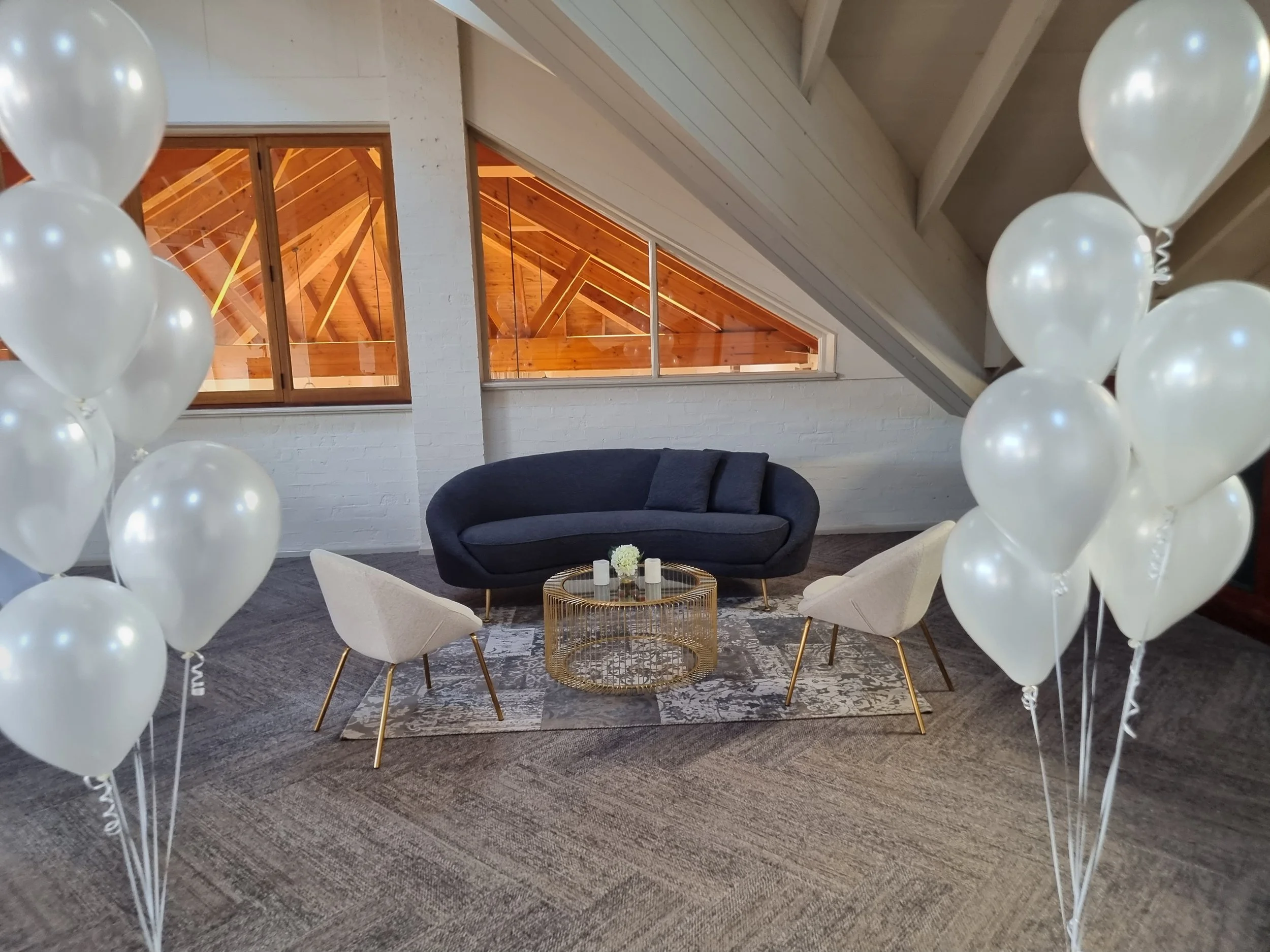 Living room with balloons framing the scene, featuring a dark blue sofa, two beige chairs with gold legs, a round gold coffee table, a vase with flowers, on a patterned rug, under a sloped ceiling with wooden beams, and windows showing a wooden attic