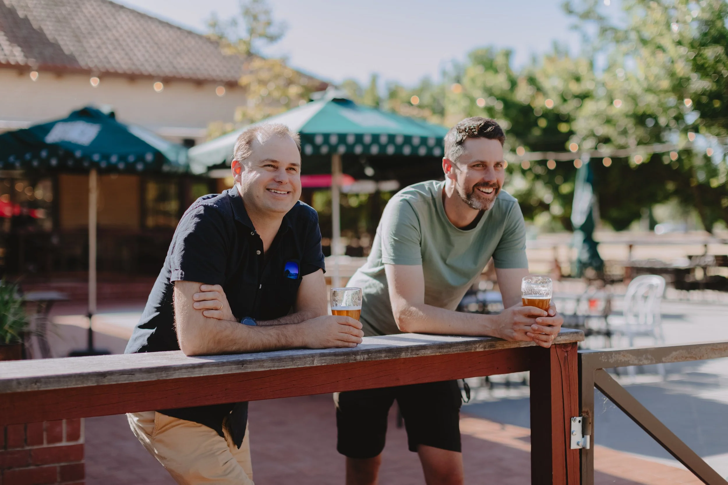 Two men smiling and holding glasses of beer at an outdoor bar or patio on a sunny day, with green umbrellas and trees in the background.