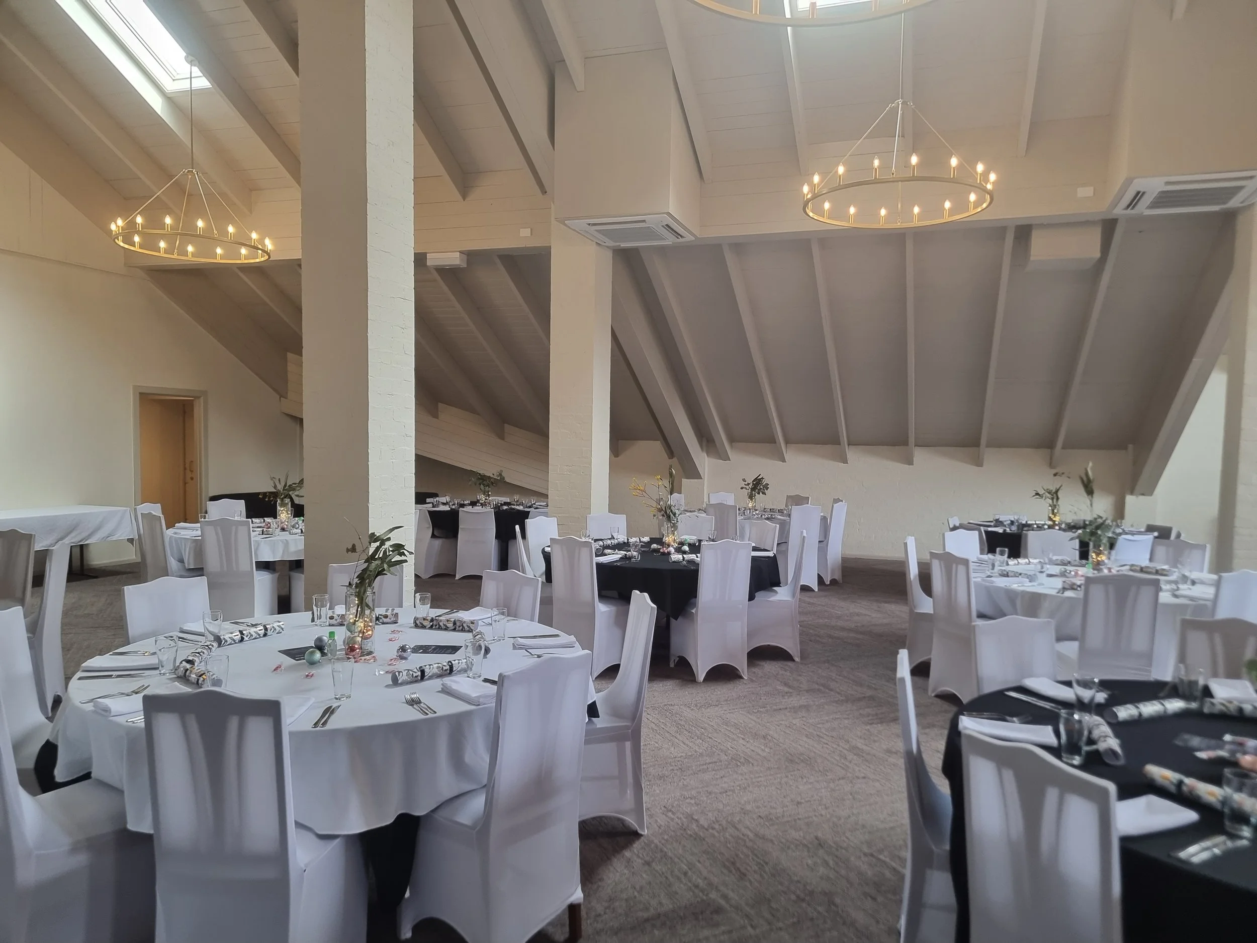 Elegant event space with round tables covered in black and white tablecloths, decorated with vases of greenery, set with glasses, silverware, and napkins, inside a spacious room with high vaulted ceilings, pendant chandeliers, and beige carpeted floo