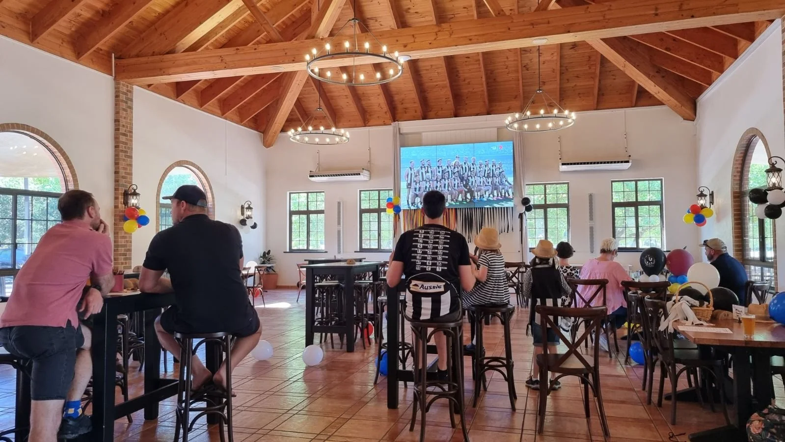 People watching a sports game on a large screen in a decorated room with balloons, tables, and chairs.