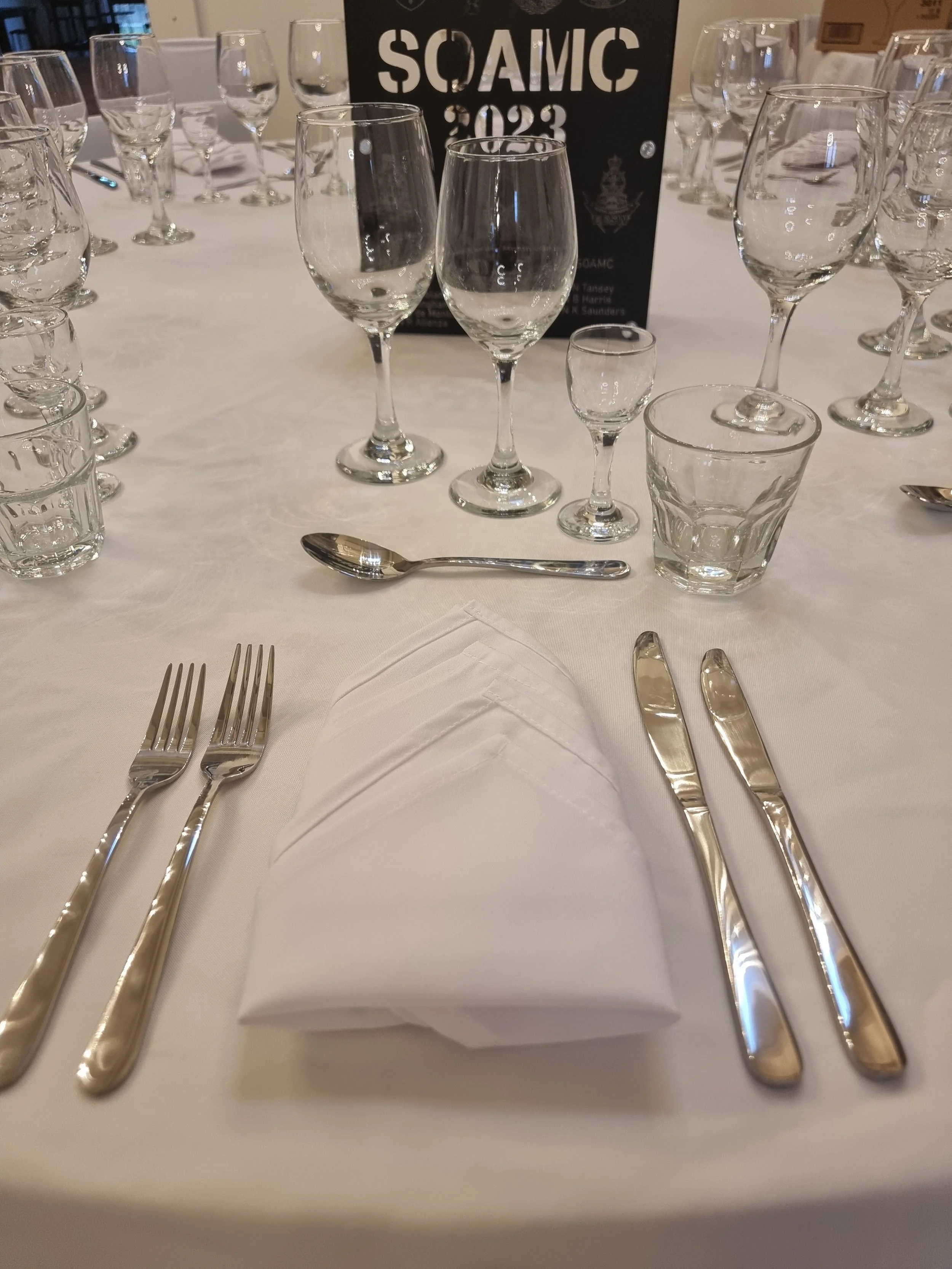 An elegant banquet table setup with white tablecloth, neatly folded napkin, silverware including two forks on the left and two knives on the right, a spoon above the napkin, and various wine glasses. In the background, a sign reads "SOAMIC 2023."