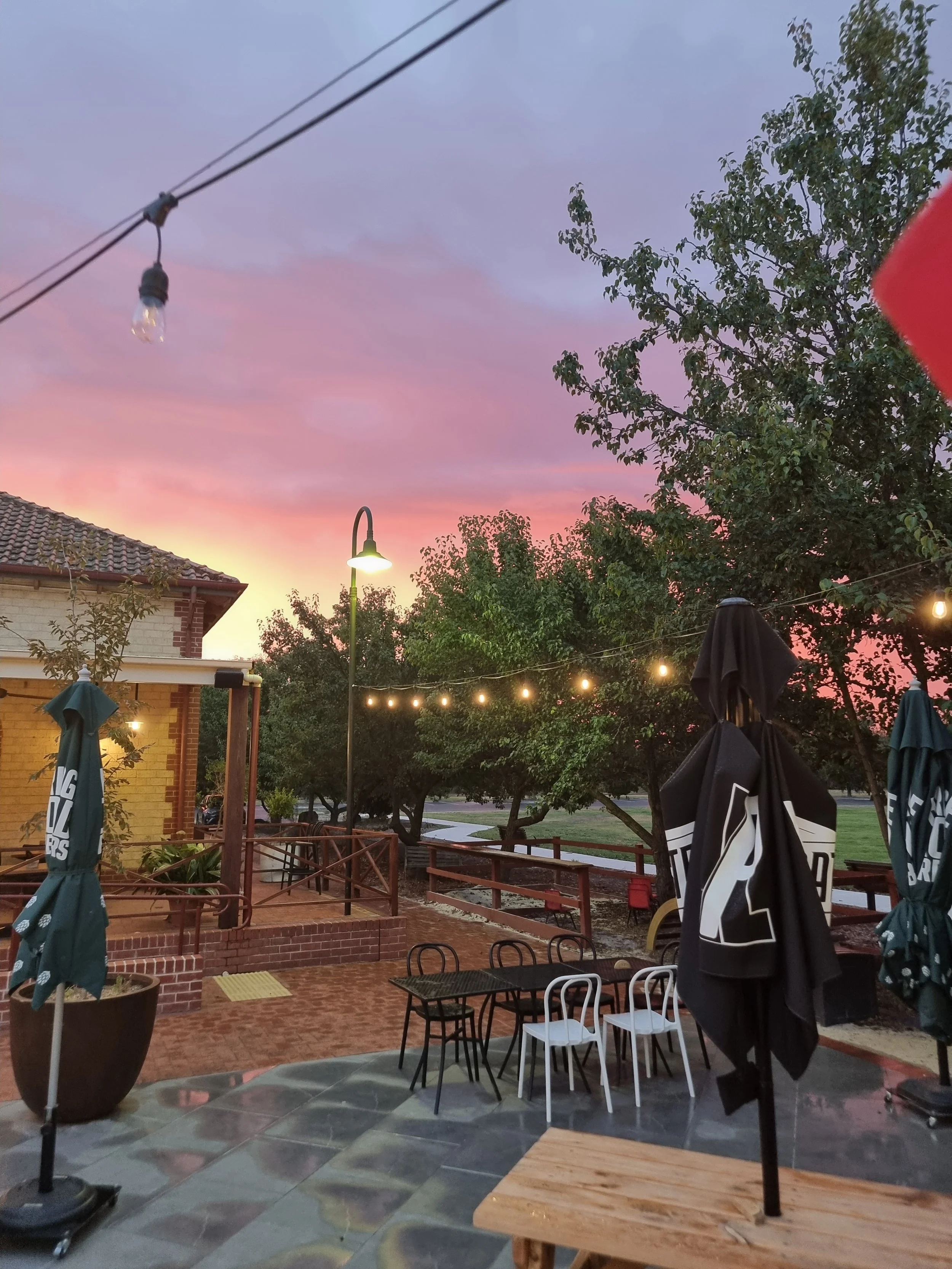 An outdoor patio area at sunset with string lights, trees, and chairs, under a sky with pink and purple clouds.
