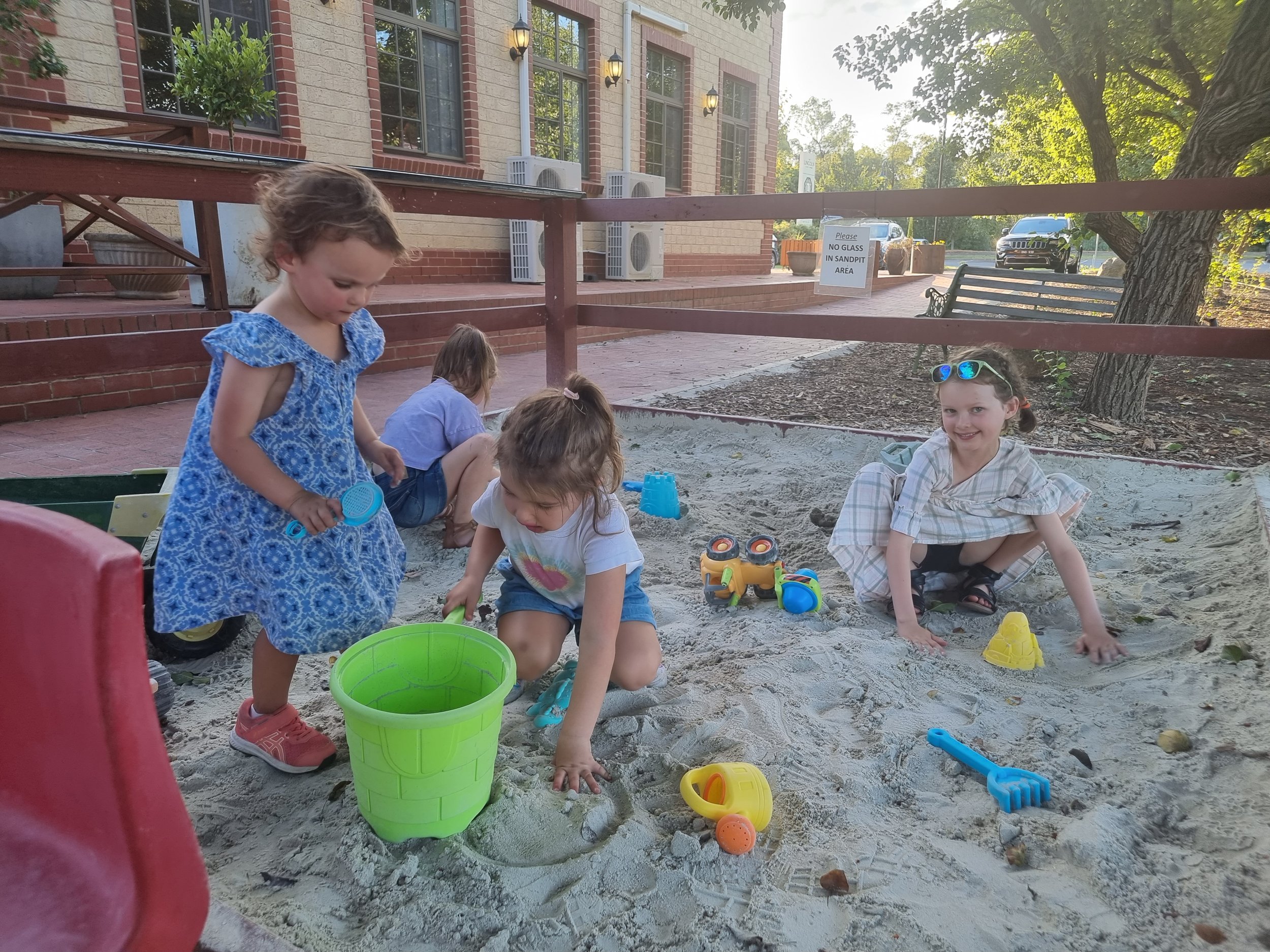 Four young girls playing in a sandbox with toys, some sitting and some standing, outside near a building and trees.