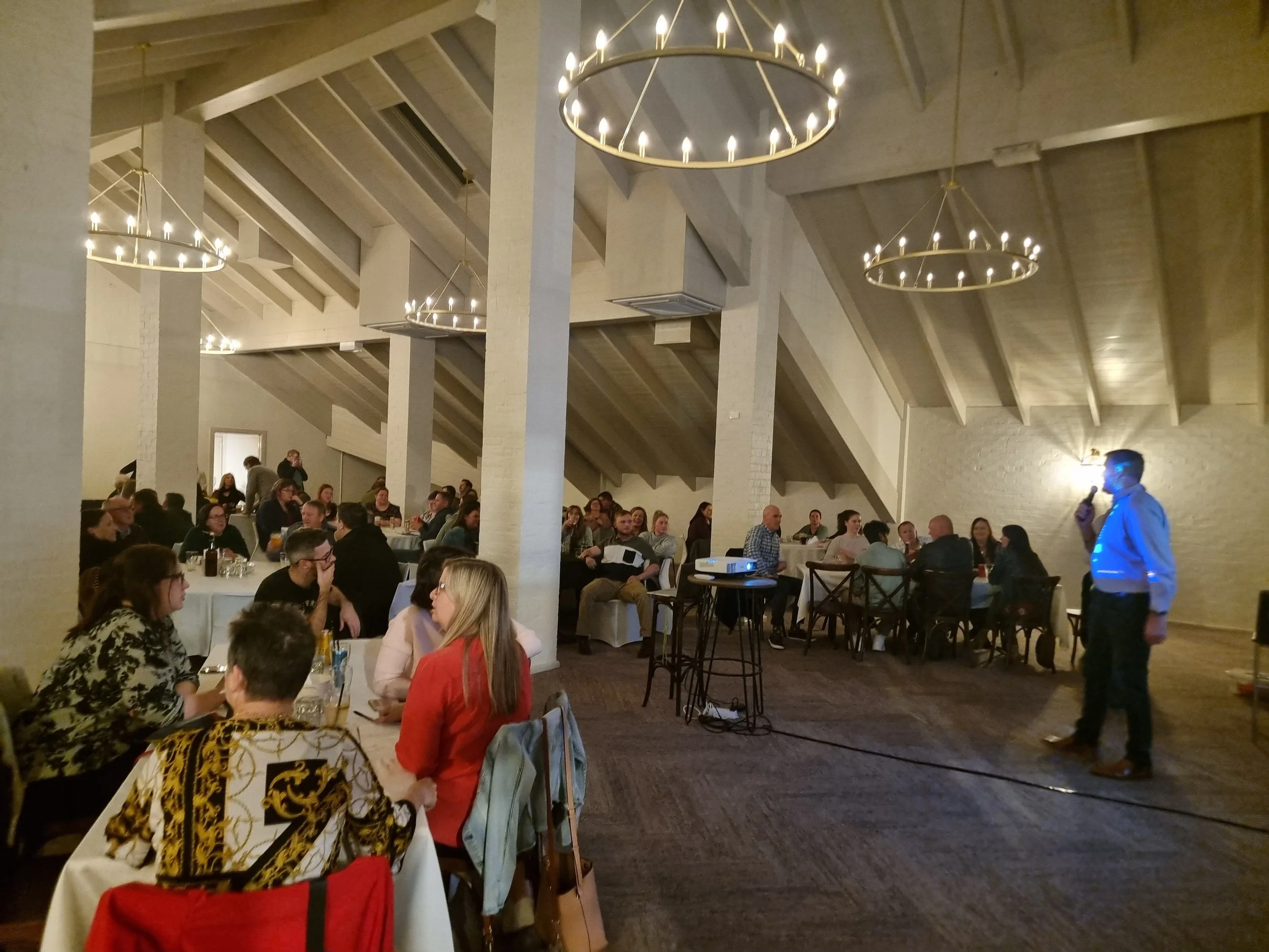 Indoor event with people gathered at tables, a speaker standing with a microphone, and circular chandeliers hanging from the ceiling