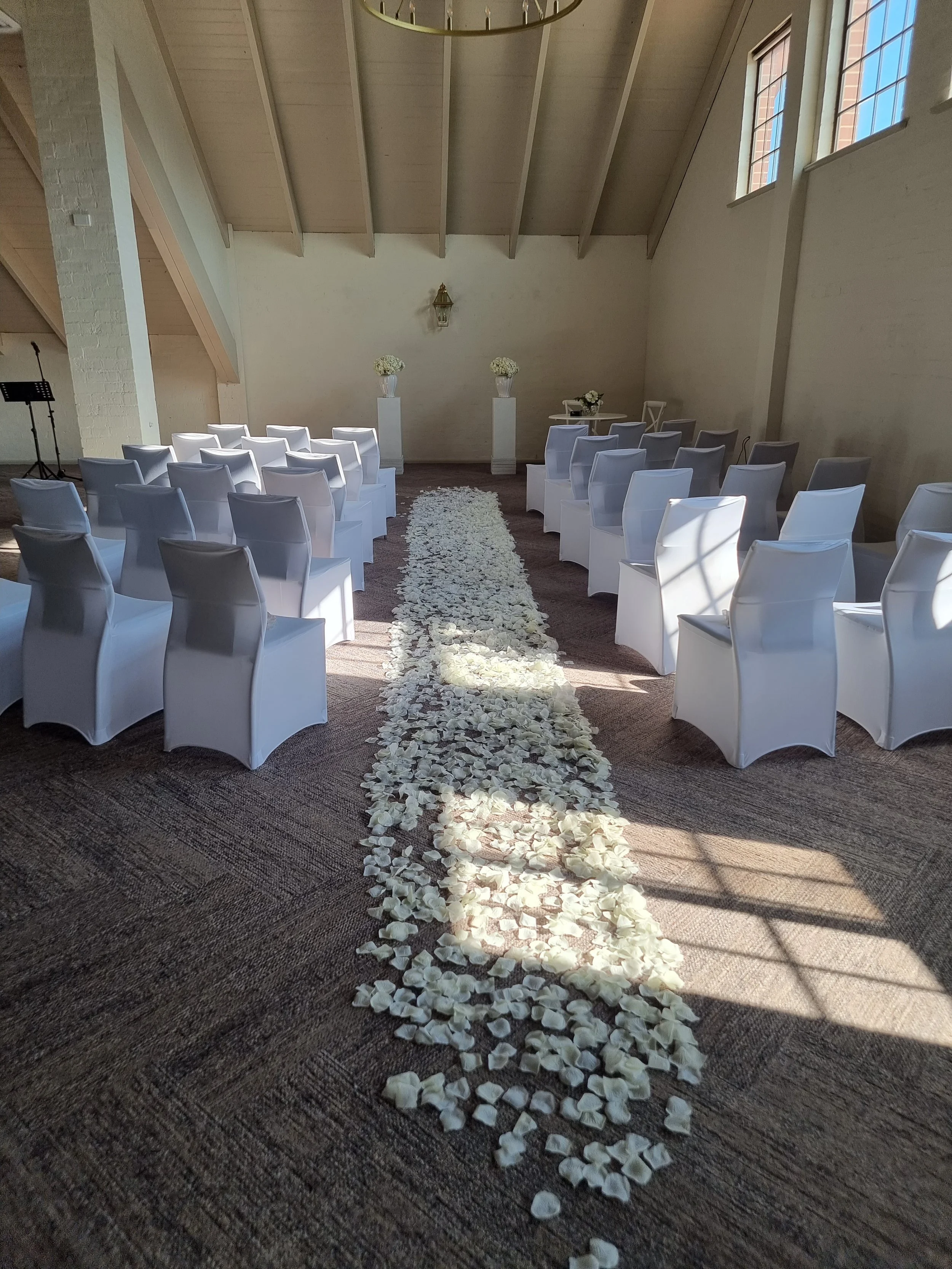 Wedding ceremony setup with white chairs arranged on both sides of a flower petal aisle leading to an altar with flowers and decorative vases inside a room with high wooden ceiling and windows.