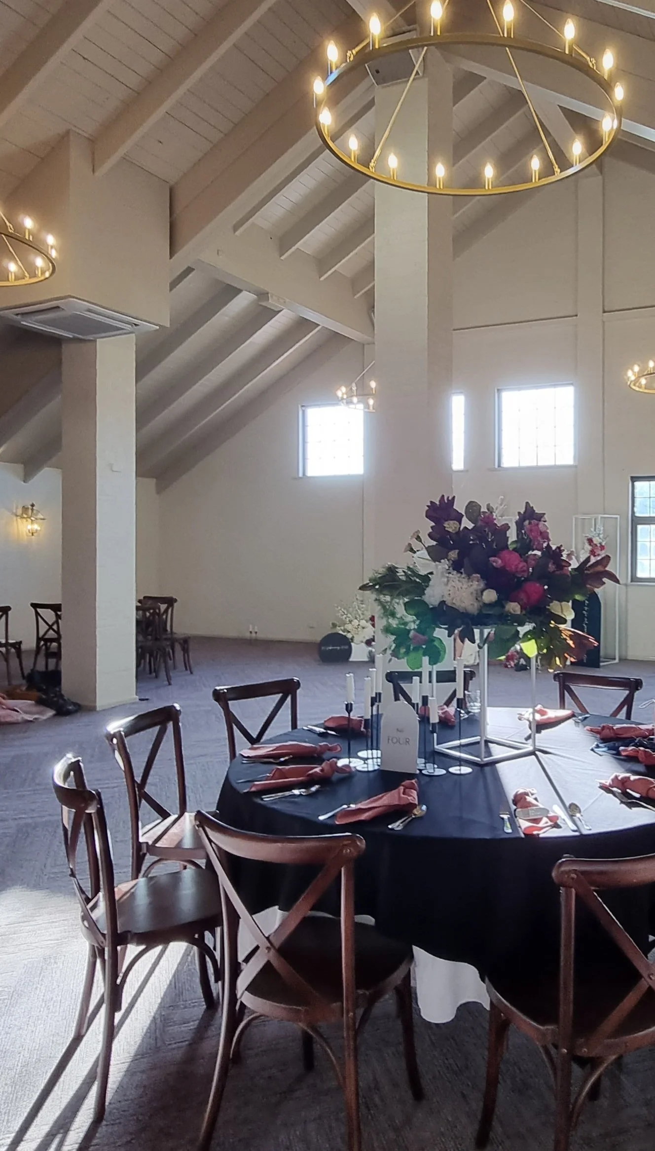 A round table with a black tablecloth, set with napkins, utensils, and a centerpiece of flowers, in a large event hall with high vaulted ceilings and chandeliers.