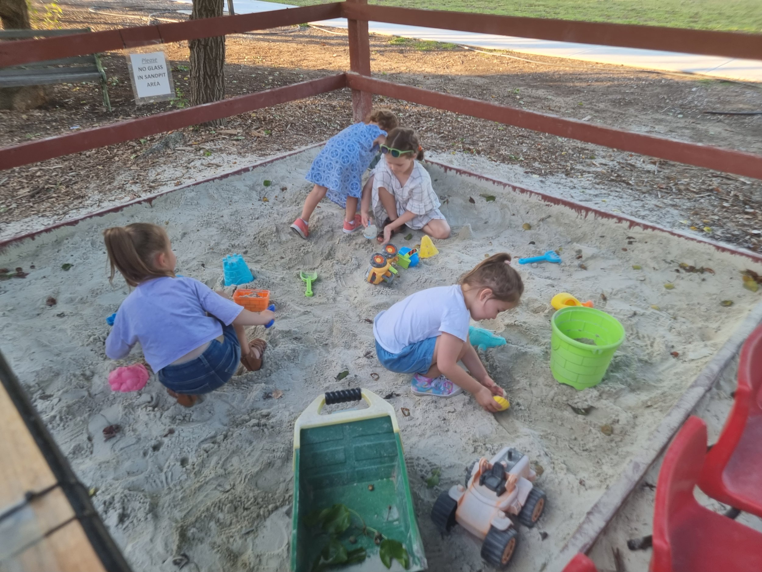 Four children playing in a sandbox surrounded by a red metal fence, with toys like shovels, buckets, and a toy vehicle, outdoors in a park or playground area.