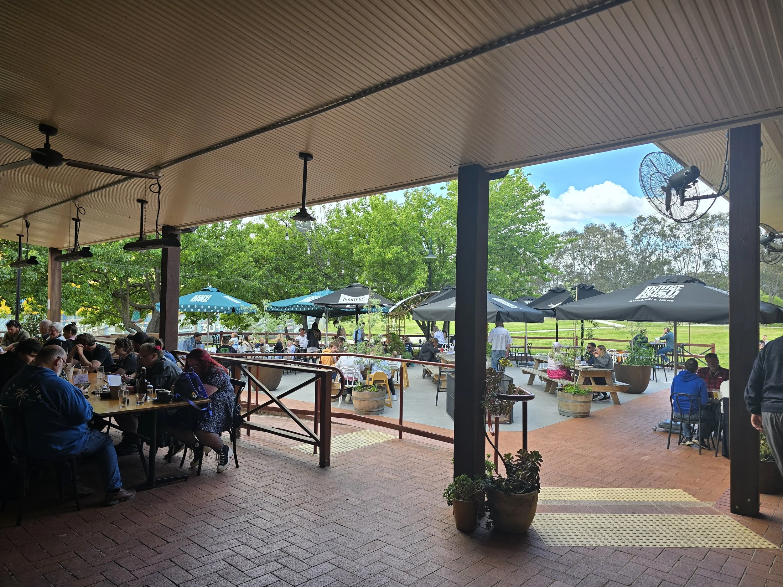 Outdoor dining area at a restaurant with people sitting at tables under umbrellas, trees and a grassy field in the background, and fans mounted on the roof.