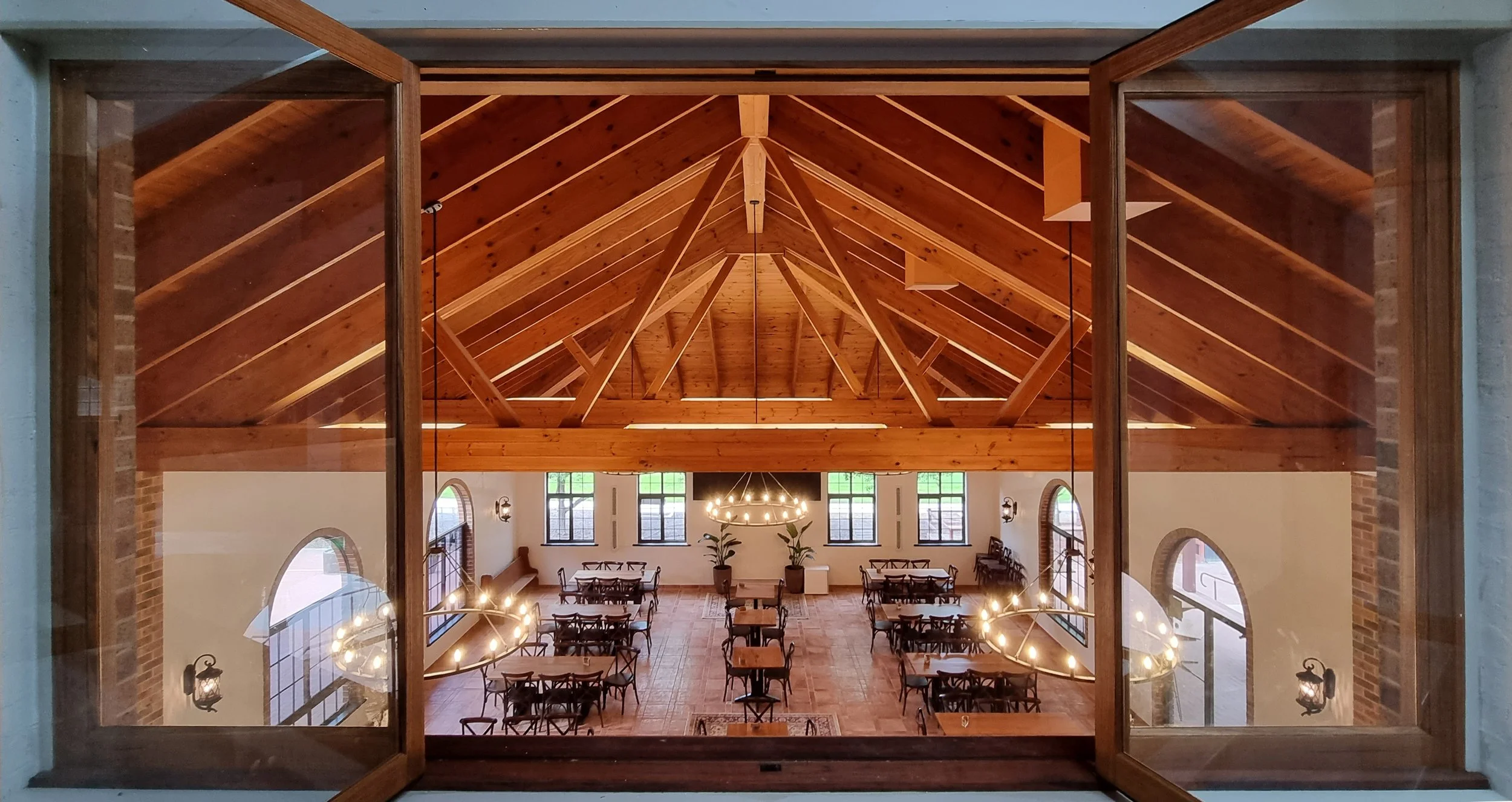 View of a spacious indoor dining area with wooden vaulted ceiling, large windows, and chandeliers, seen through an open window.