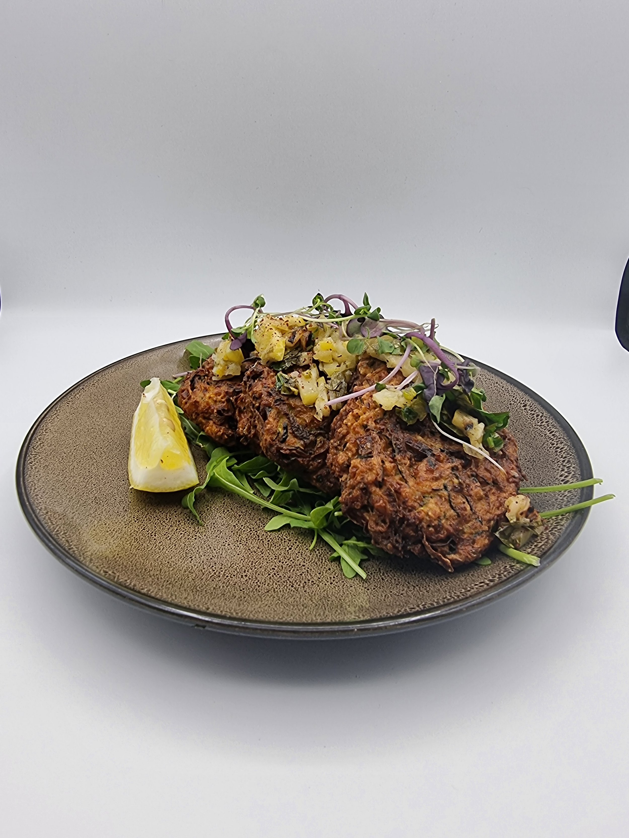 Plate with three pieces of cooked falafel garnished with microgreens, a lemon wedge, and a small amount of salad on a brown plate.