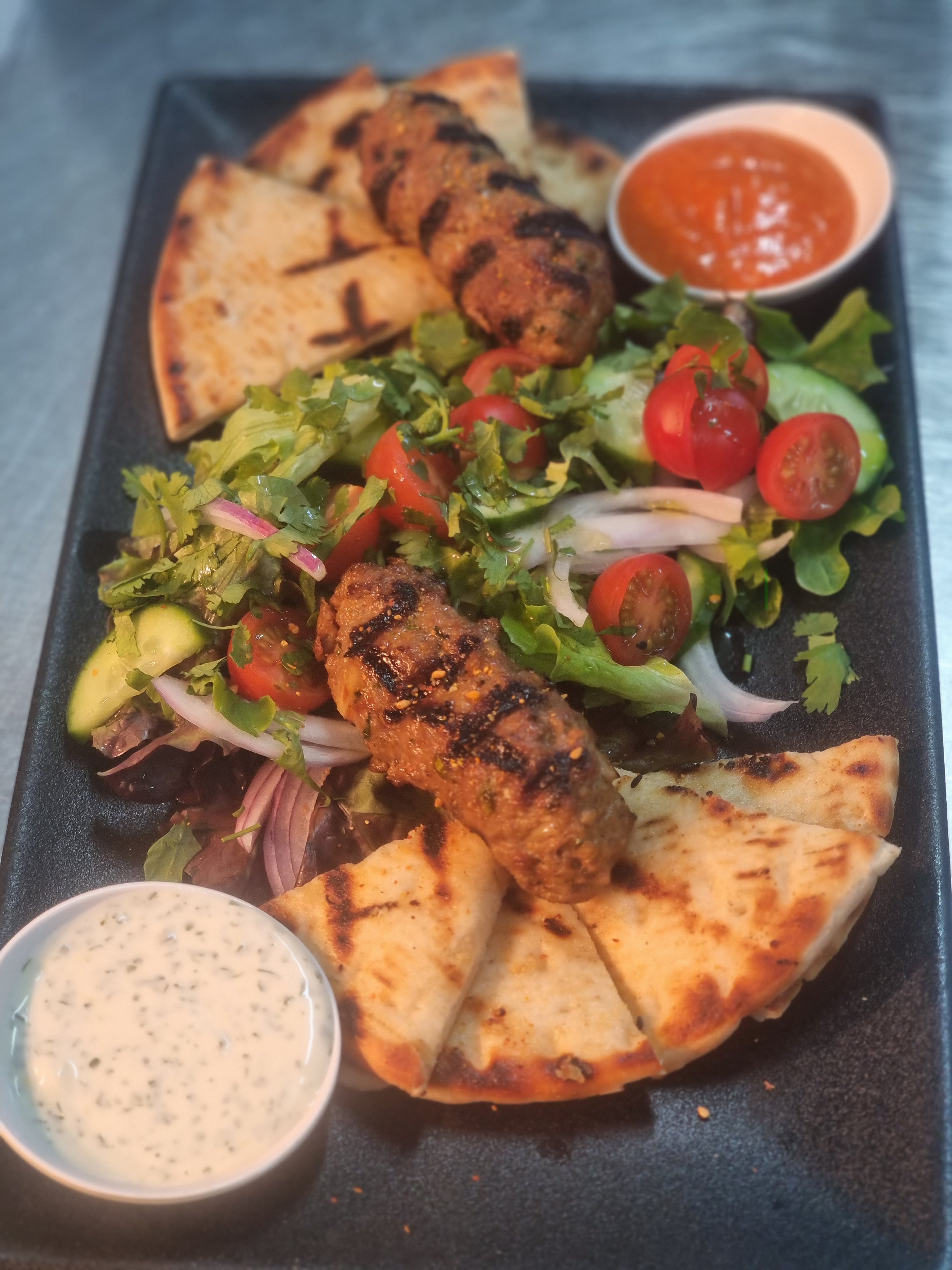 Plate of Indian food including grilled meats, naan bread, fresh salad with cherry tomatoes and cucumbers, and two small bowls of dipping sauces.