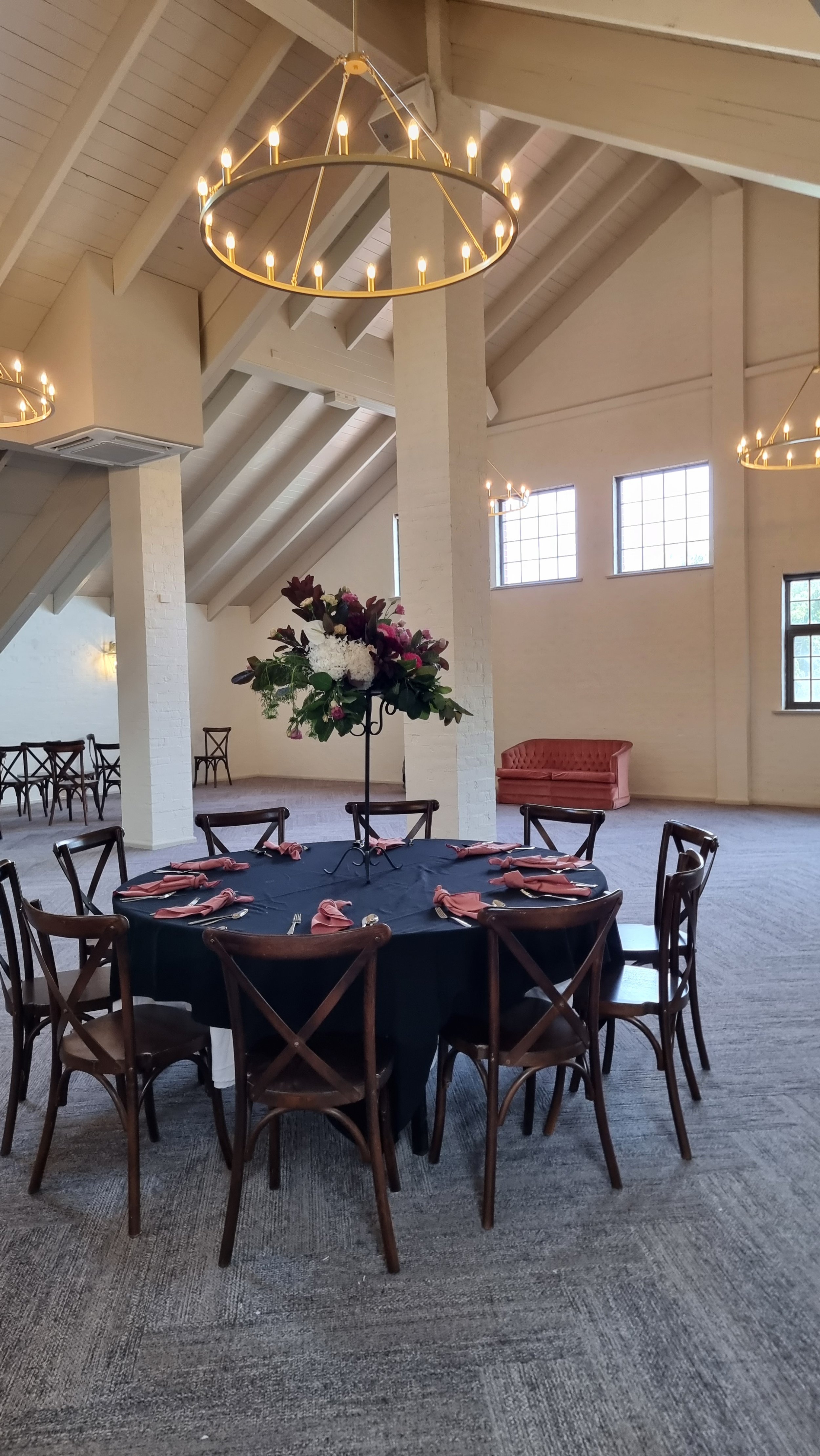 Elegant event space with a round table set with pink napkins, a floral centerpiece, and surrounded by wooden chairs. The room features high ceilings with white beams, large windows, warm lighting from chandeliers, and minimal decor.