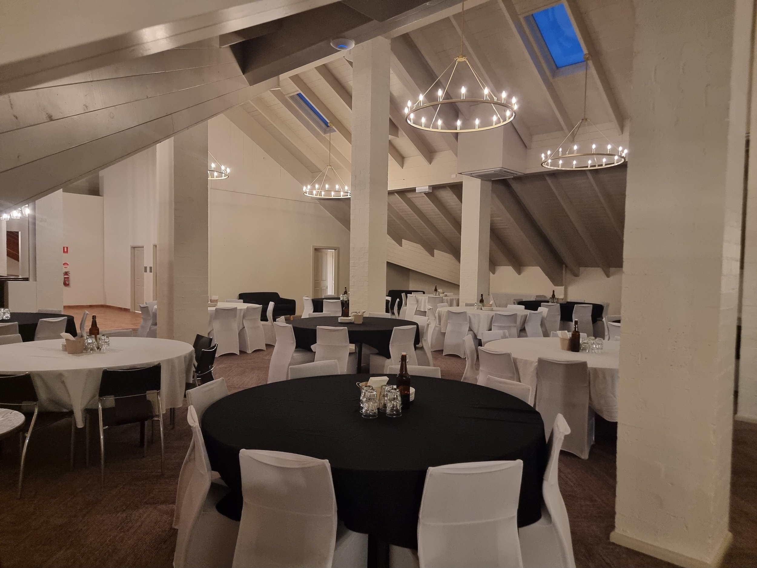 Interior of a banquet hall with round tables covered in black and white tablecloths and white chairs, chandeliers hanging from the ceiling, and a few bottles and glassware on the tables.