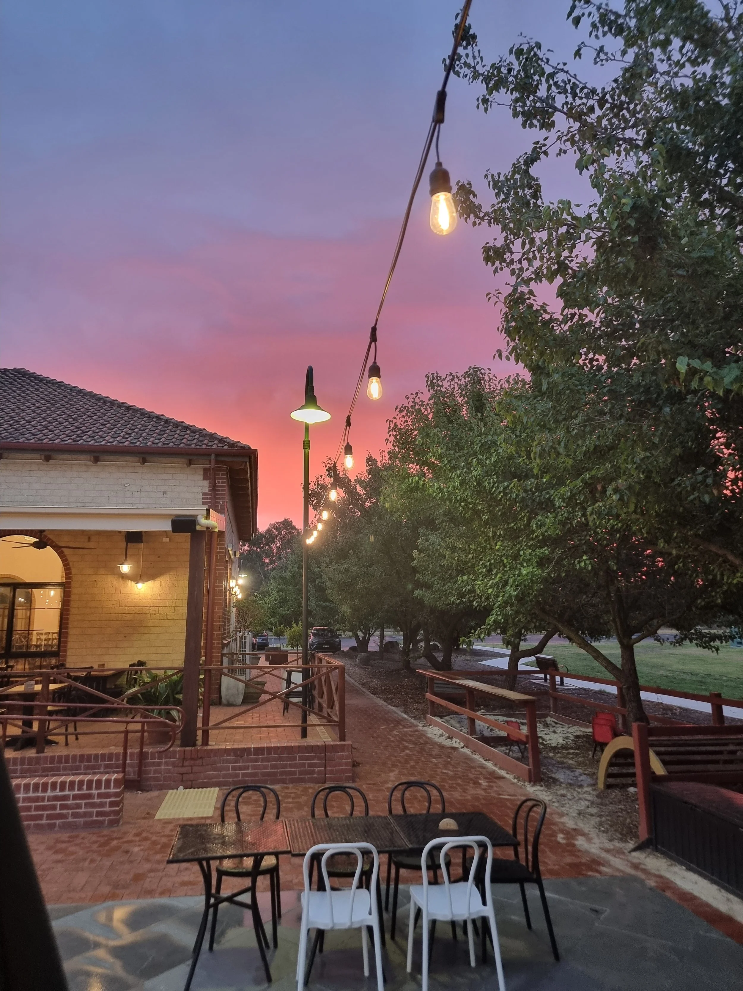 Outdoor patio scene at sunset with string lights, trees, and a brick building.