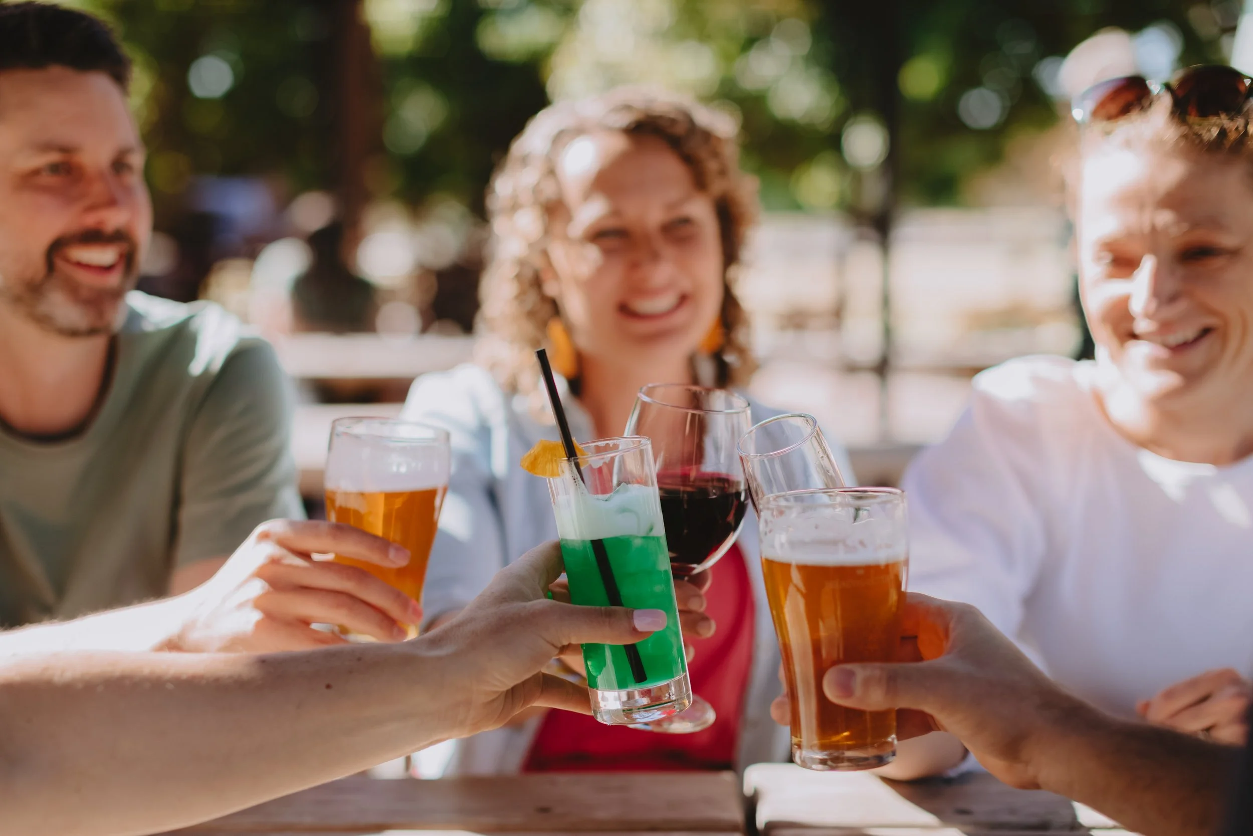 Group of friends enjoying drinks outdoors, cheersing colorful cocktails and beers at a sunny patio.