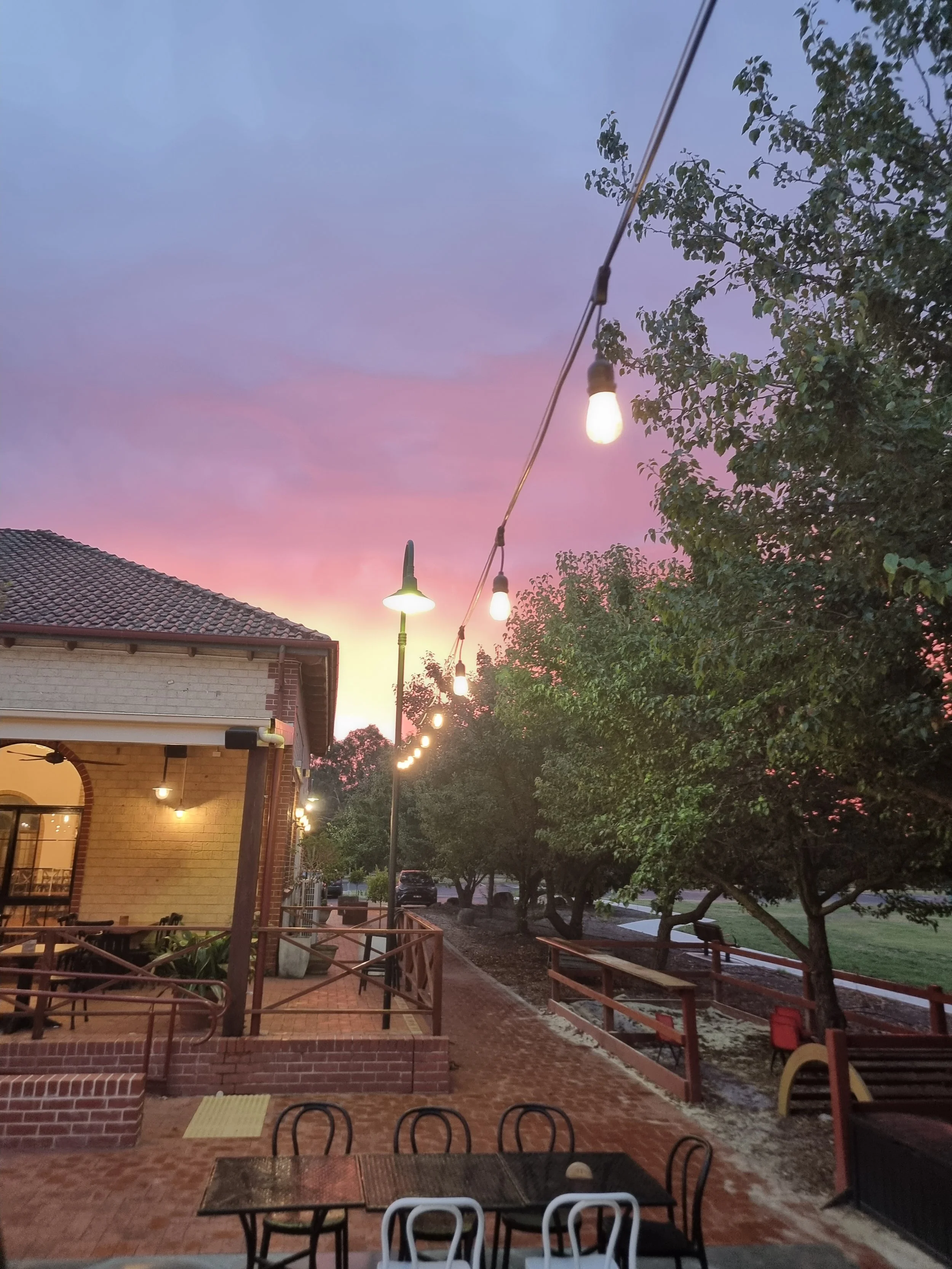Outdoor patio of a restaurant or cafe with string lights hanging, a brick building, trees, and a colorful sunset sky.