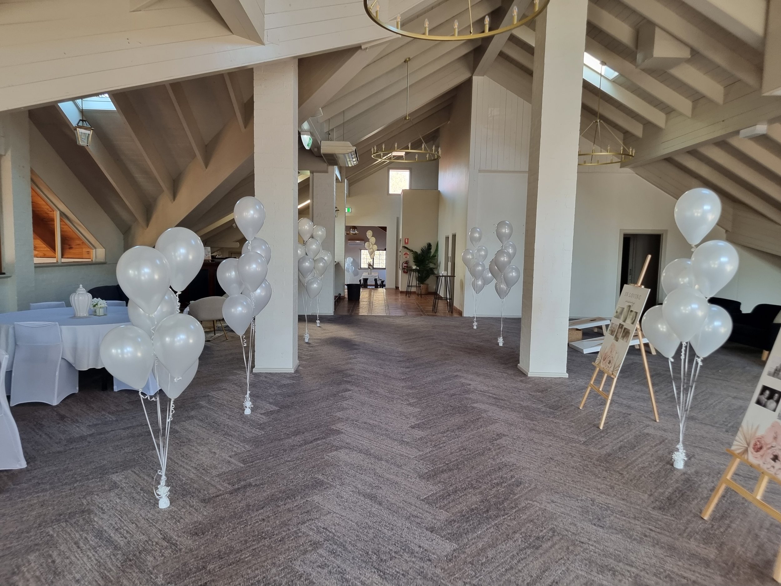 Decorated indoor event space with white balloons, tables with white tablecloths, and framed photographs on easels.