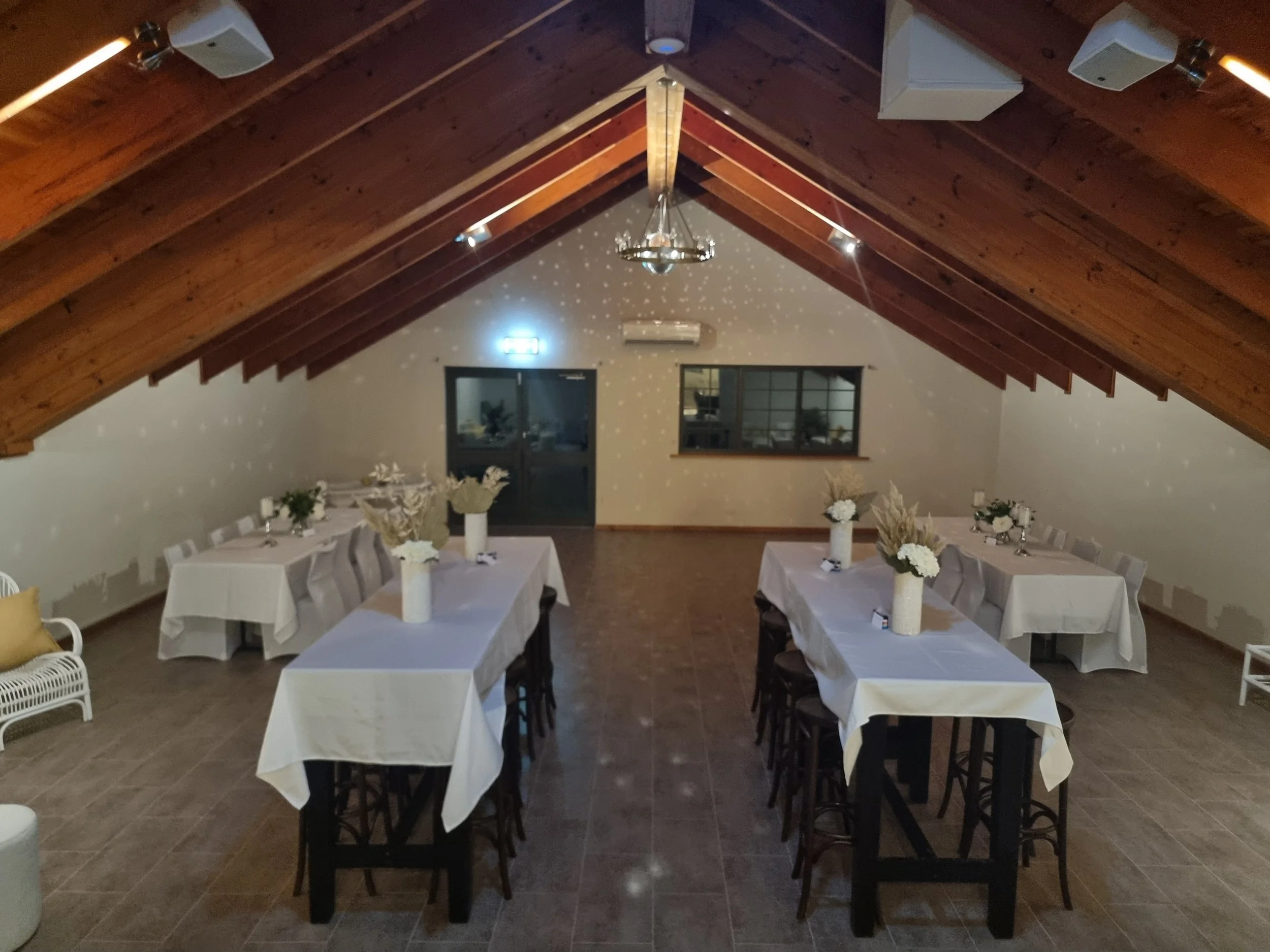 Indoor event space with sloped wooden ceiling, decorated tables with white tablecloths and floral centerpieces, and ambient lighting creating star-like patterns on the ceiling.