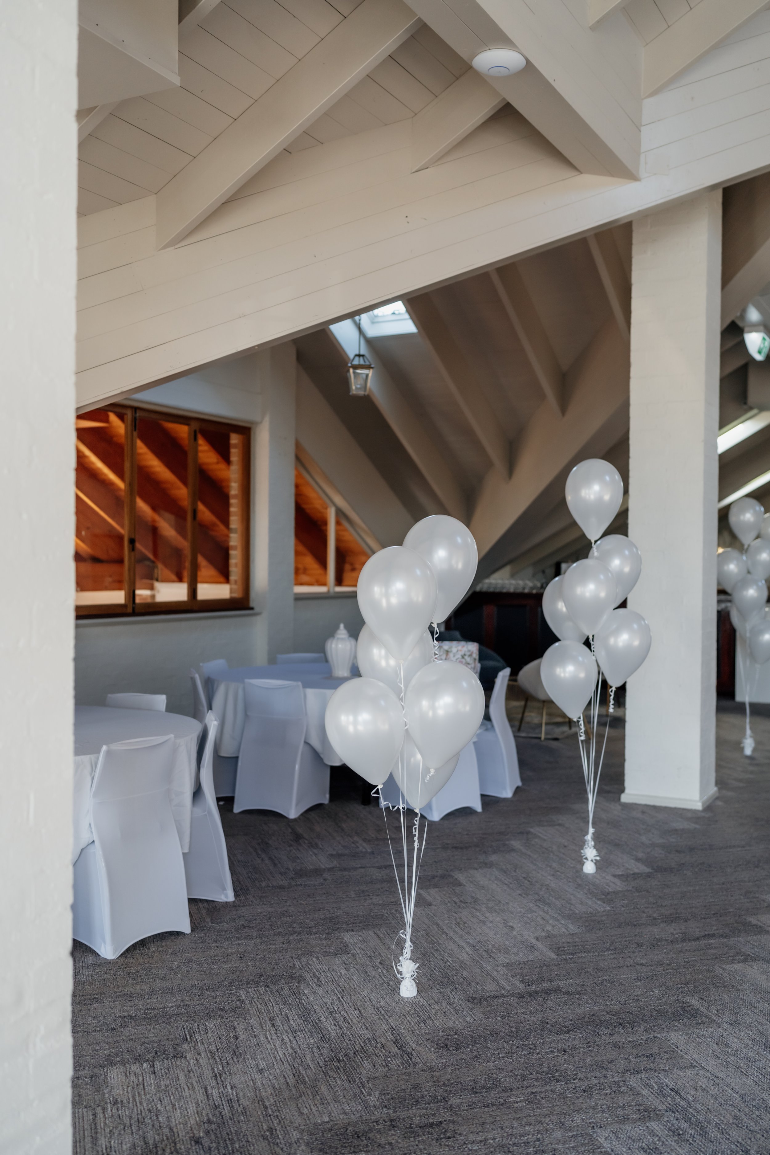 White balloons tied to the floor are arranged in front of a decorated dining area with round tables covered in white tablecloths and chairs with white covers, in an indoor space with a sloped ceiling and skylights.