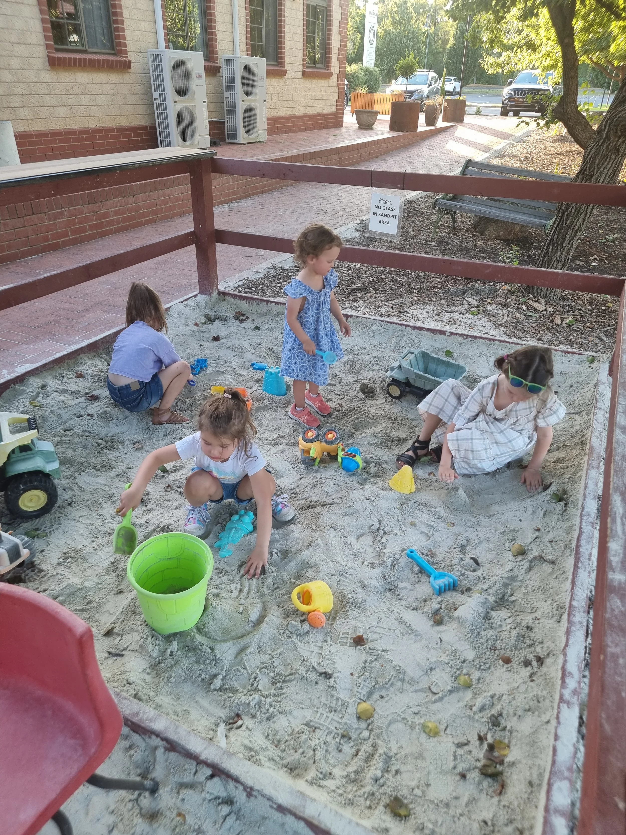 Four children playing in a sandbox at a park, surrounded by a red wooden fence, with a brick building, trees, and parked cars in the background.