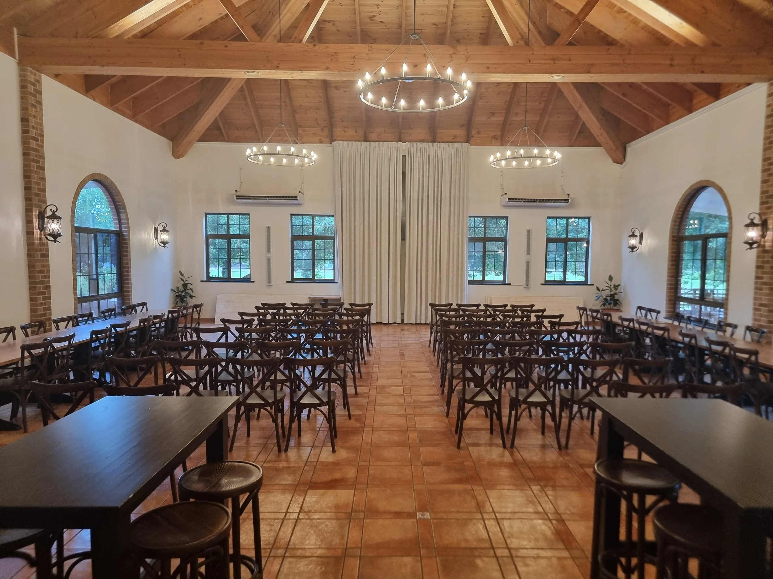 Empty banquet hall with wooden ceiling, multiple chandeliers, arched windows, tables and chairs arranged in rows, and decorative plants.