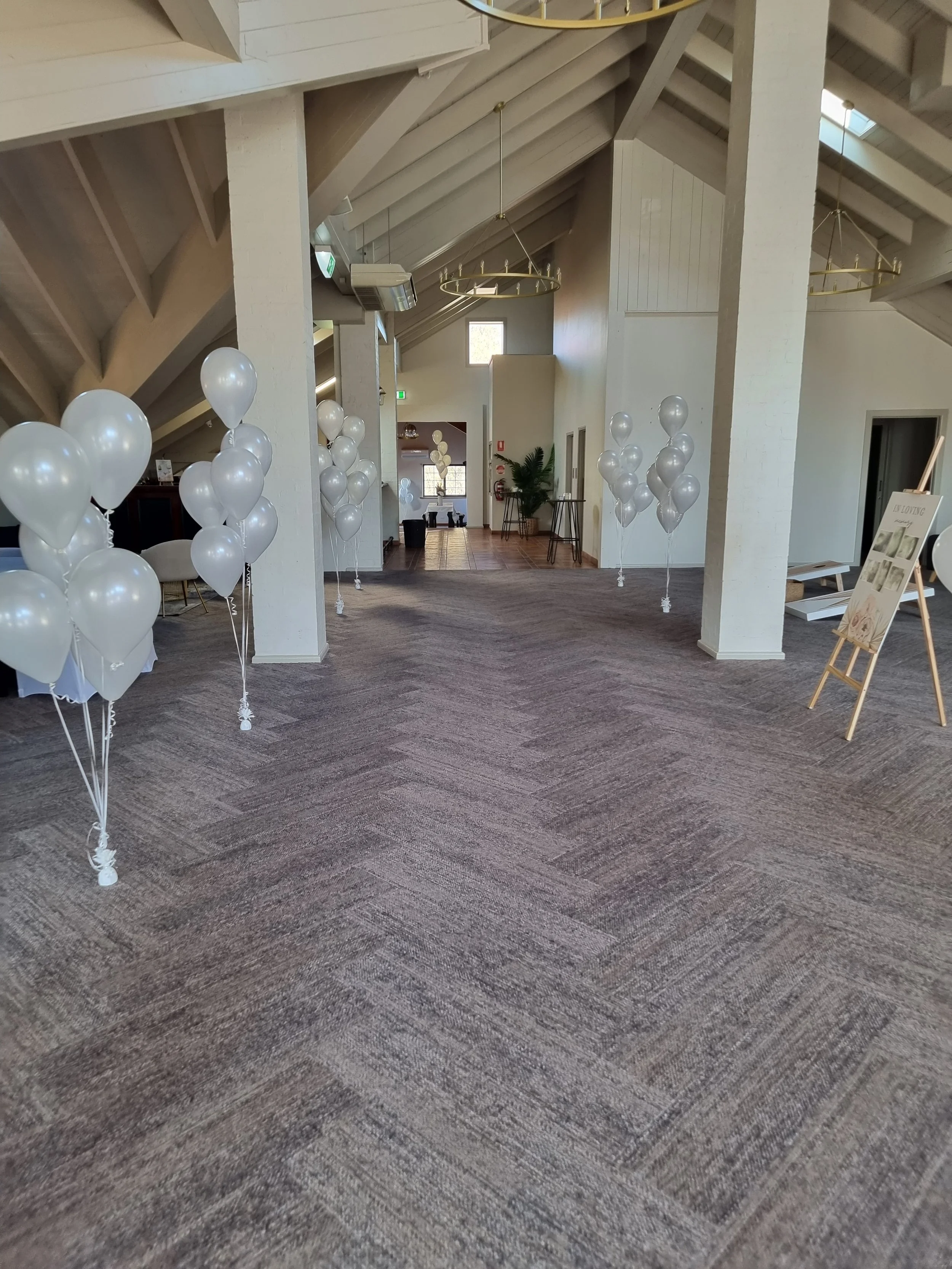 Empty event space decorated with white balloons, arranged in small clusters along the sides, with a sign on an easel to the right. The space features high, angled ceiling beams and large windows bringing in natural light.
