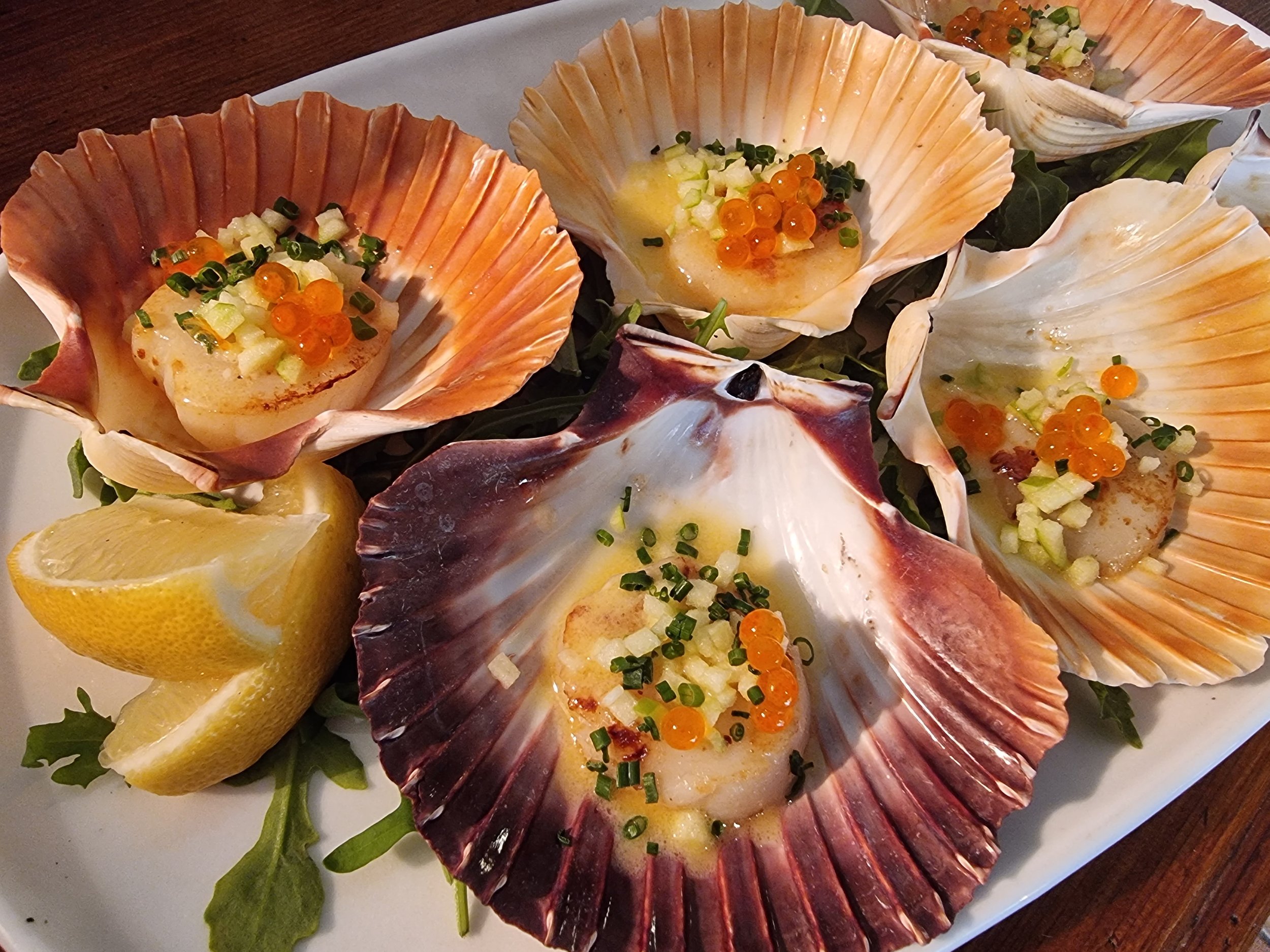 Seared scallops served in large clam shells, garnished with chopped green onions, diced cucumber, orange fish roe, and a citrus sauce, accompanied by lemon wedges.